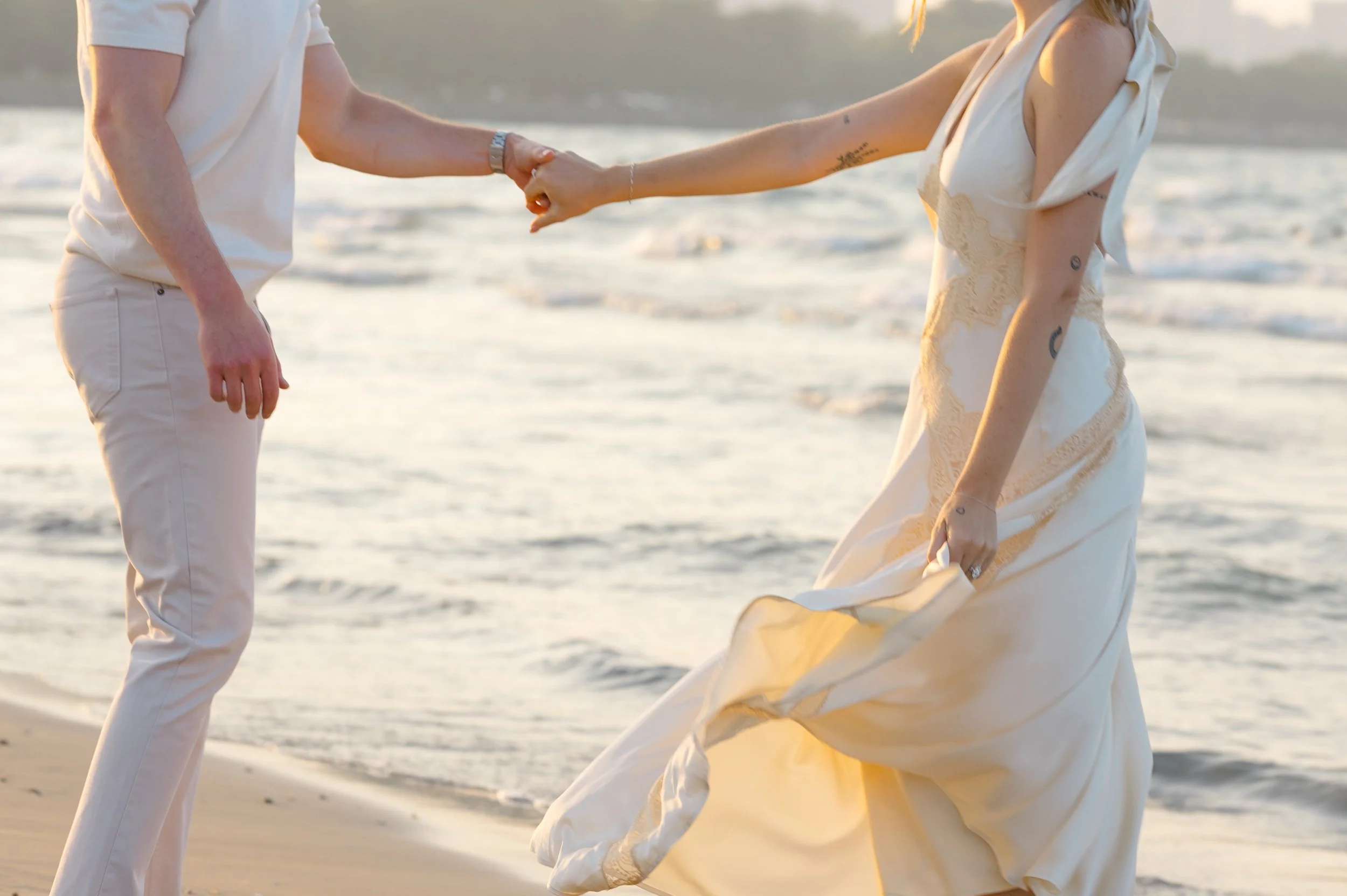 A couple holding hands at the beach during sunset, with the woman wearing a light-colored dress and the man dressed in light-colored attire, with the ocean and waves in the background.