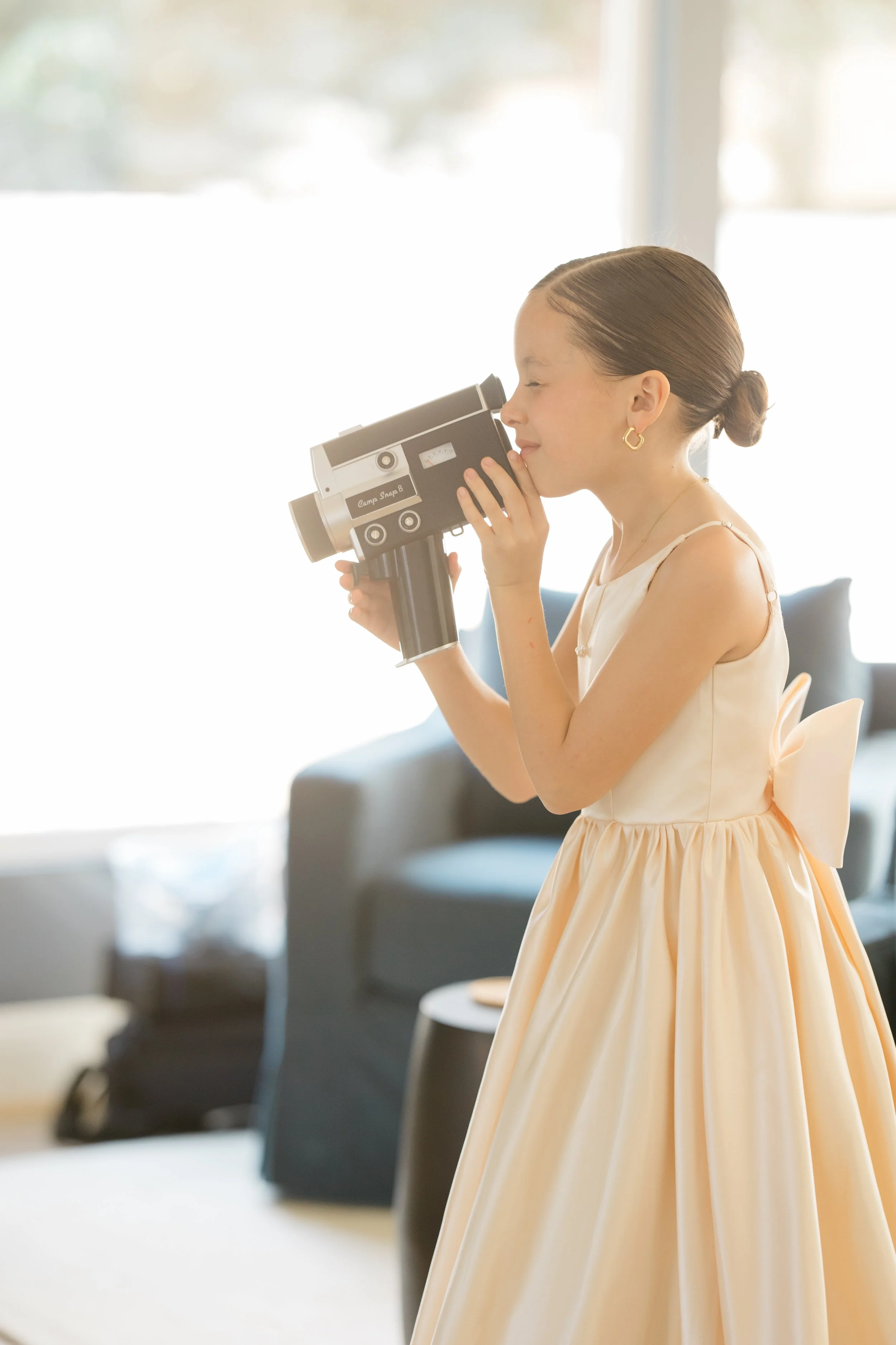A young girl in a peach-colored dress with a large bow on the back is using a telescope indoors. The background is bright with a large window and blurred furniture.