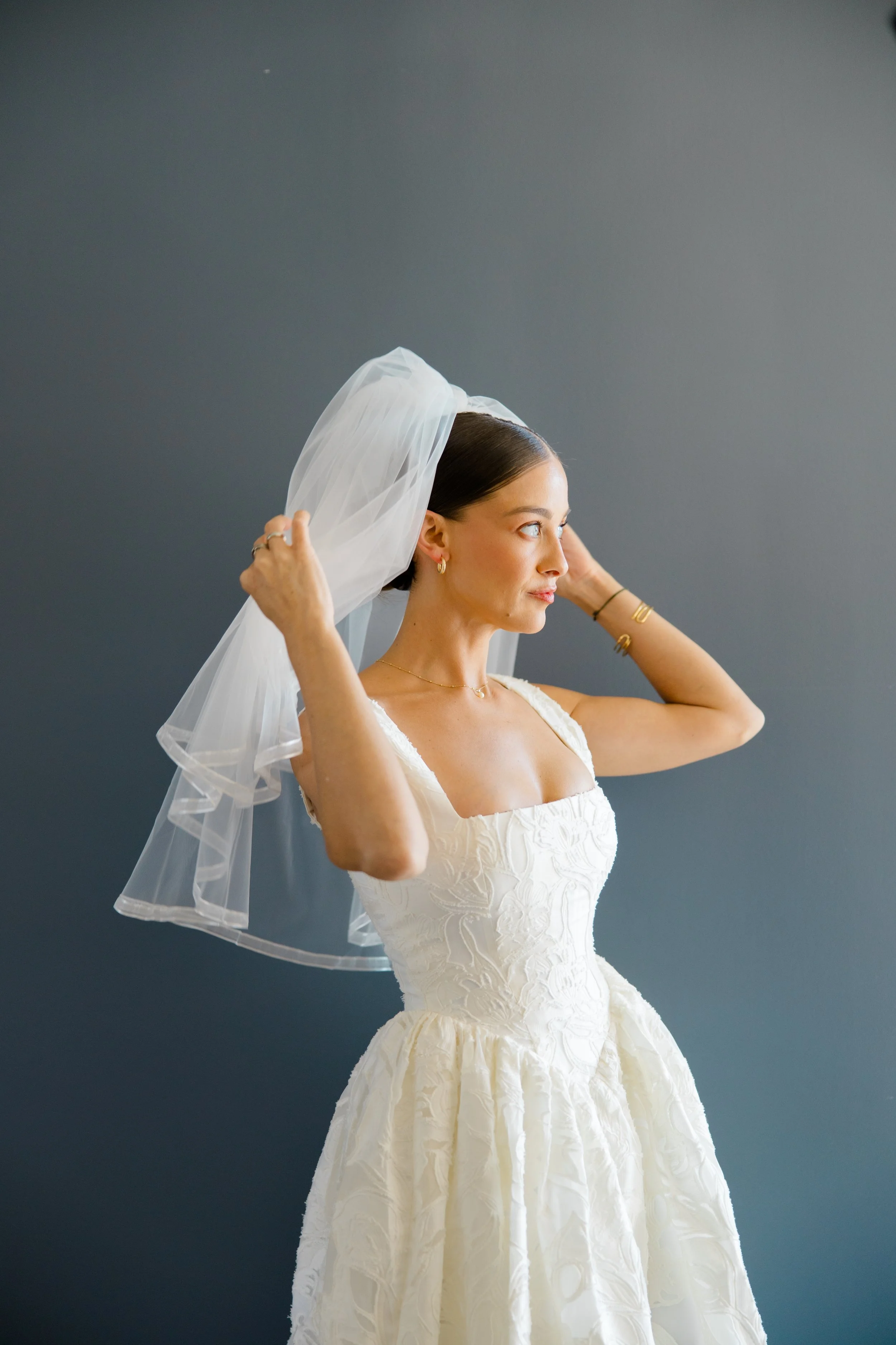 A woman in a white wedding dress putting on a veil against a dark gray background.