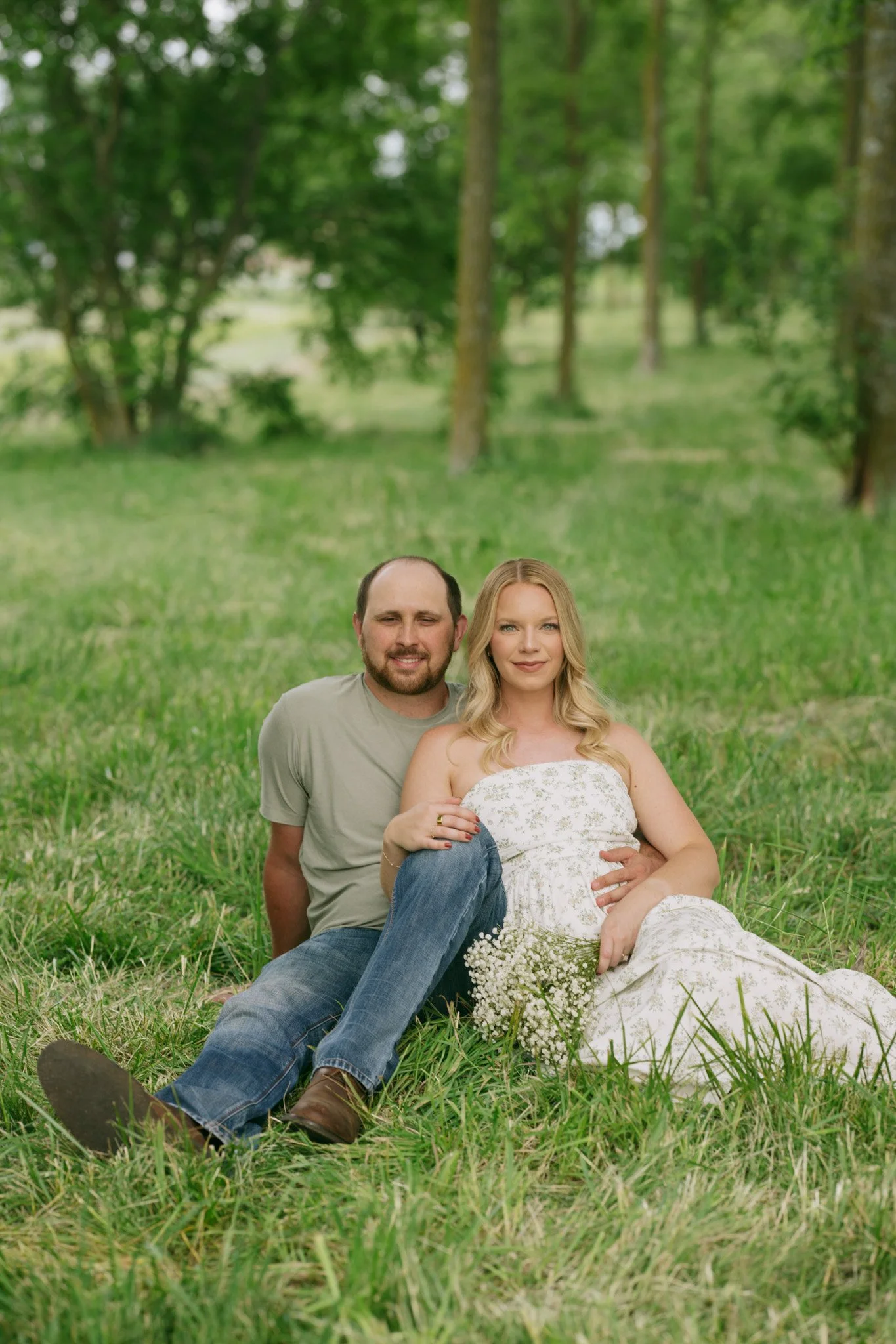 A happy couple sitting on the grass in a park, with trees in the background.