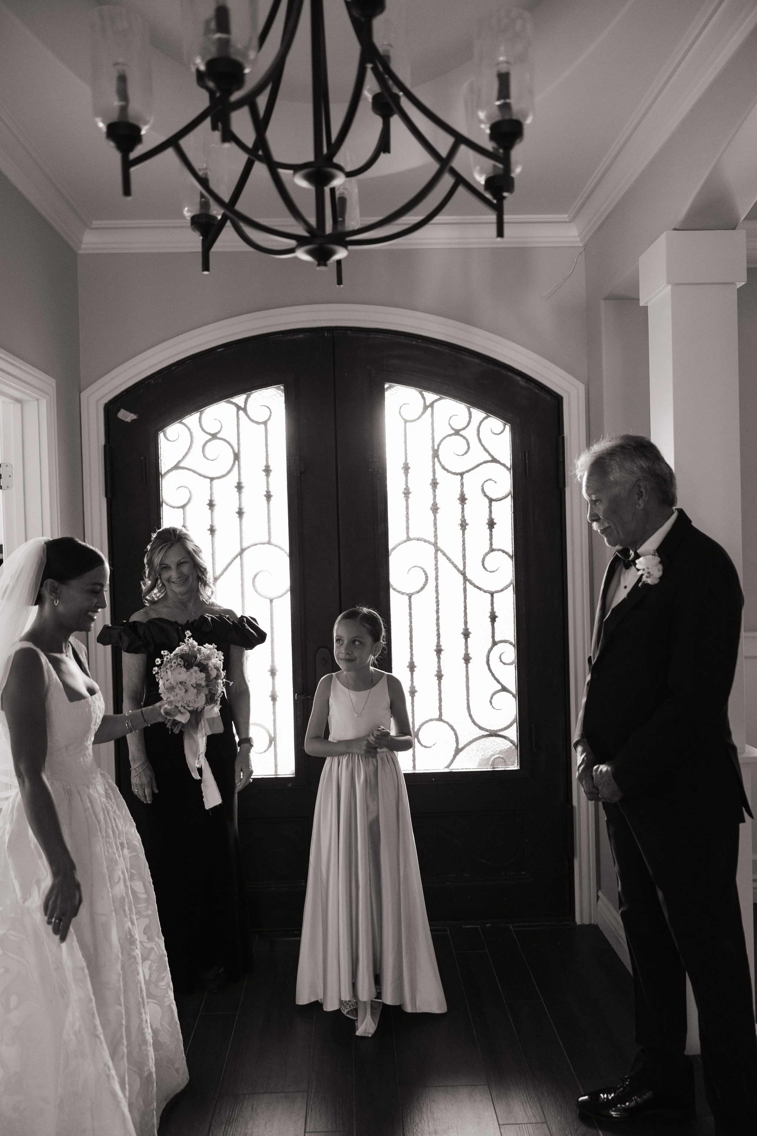 A group of people, including a bride, a young girl, and an older man, gathered in front of a decorative door with ironwork. The scene appears to be at a wedding celebration.