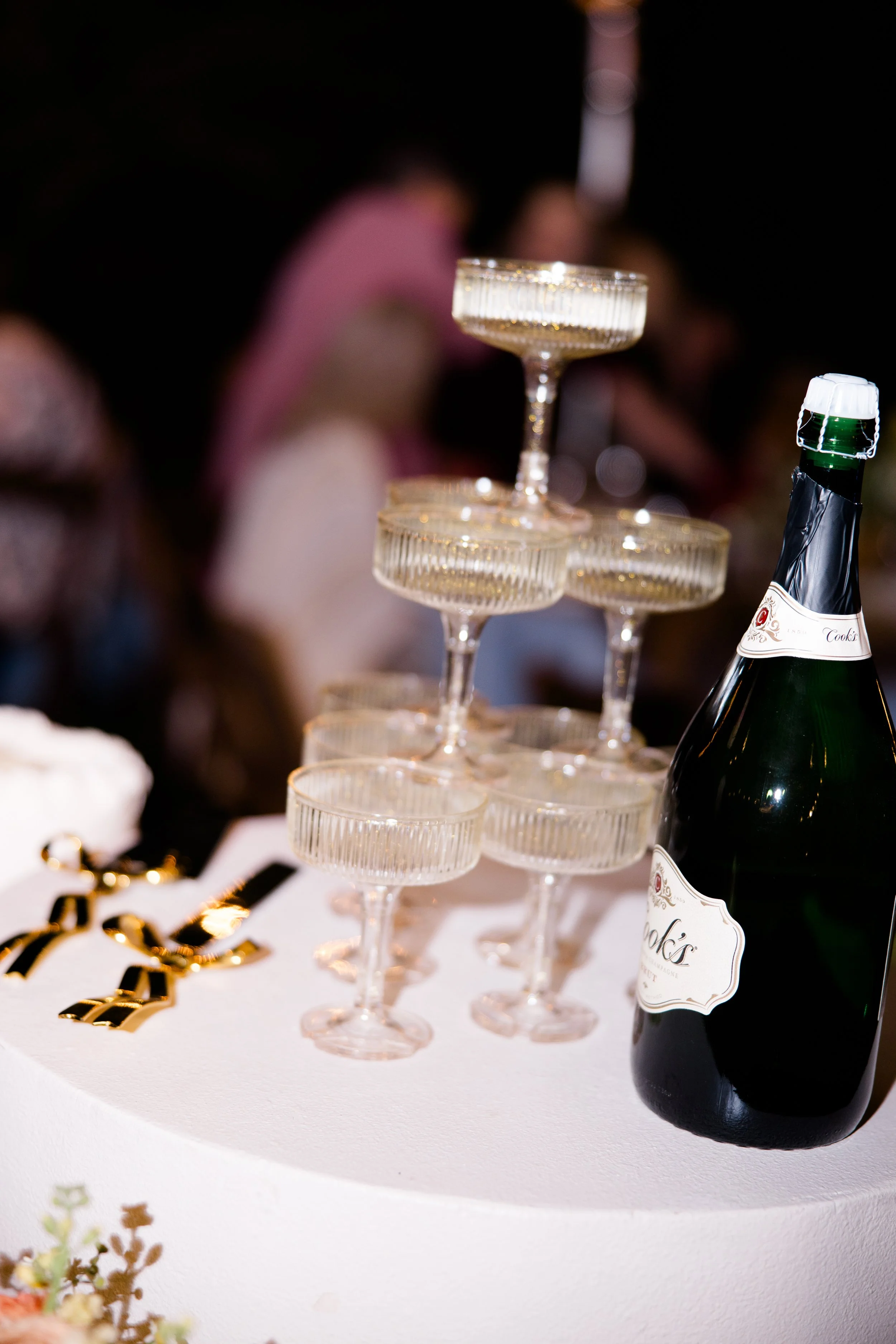 A champagne bottle with gold foil and white label placed on a white table decorated with gold confetti, with a tower of empty champagne glasses stacked using the base of each glass as the top of the next.