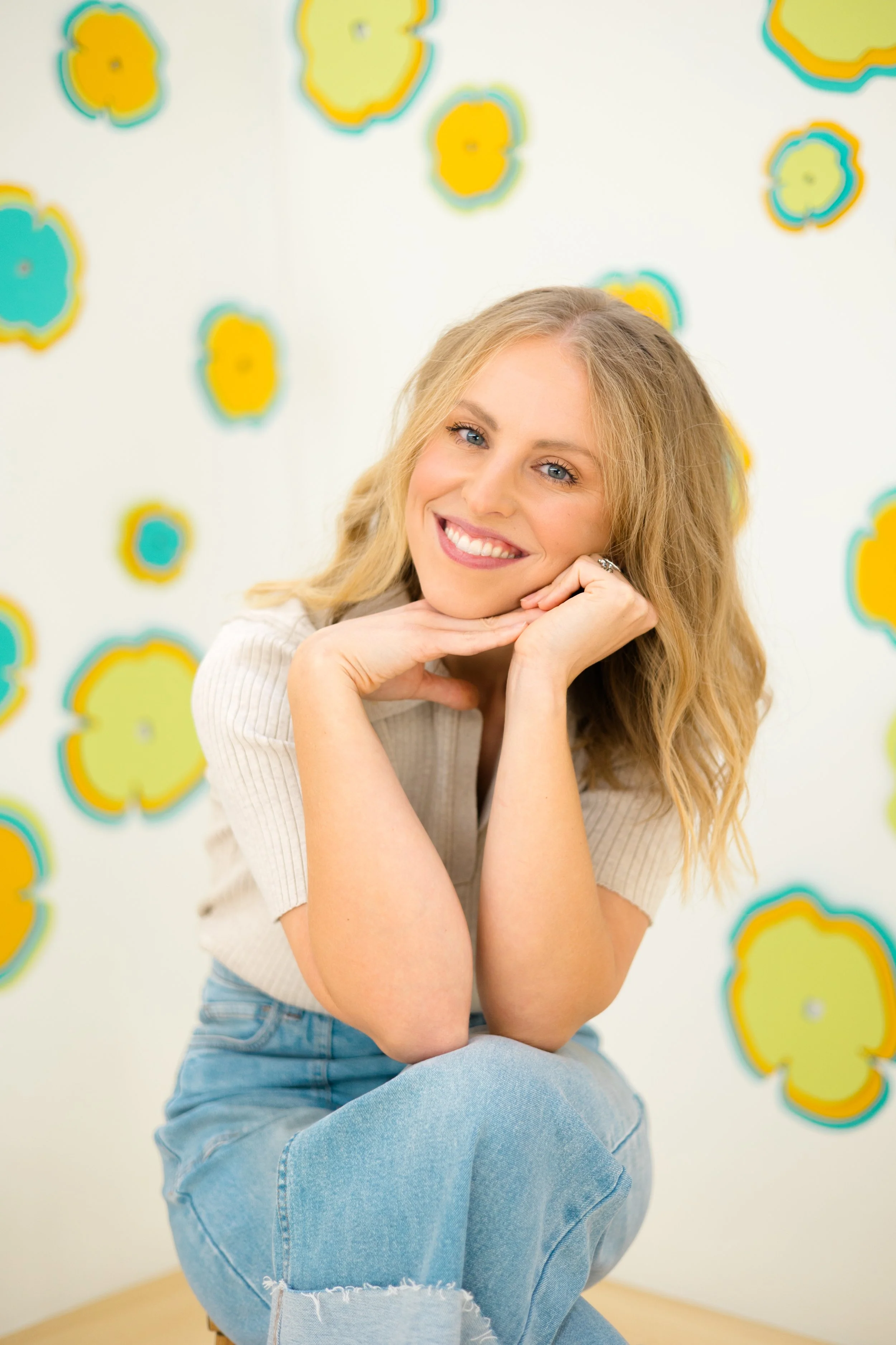 A woman with long blonde hair, blue eyes, and a bright smile resting her chin on her hand, sitting in front of a colorful abstract background with yellow, blue, and orange shapes.
