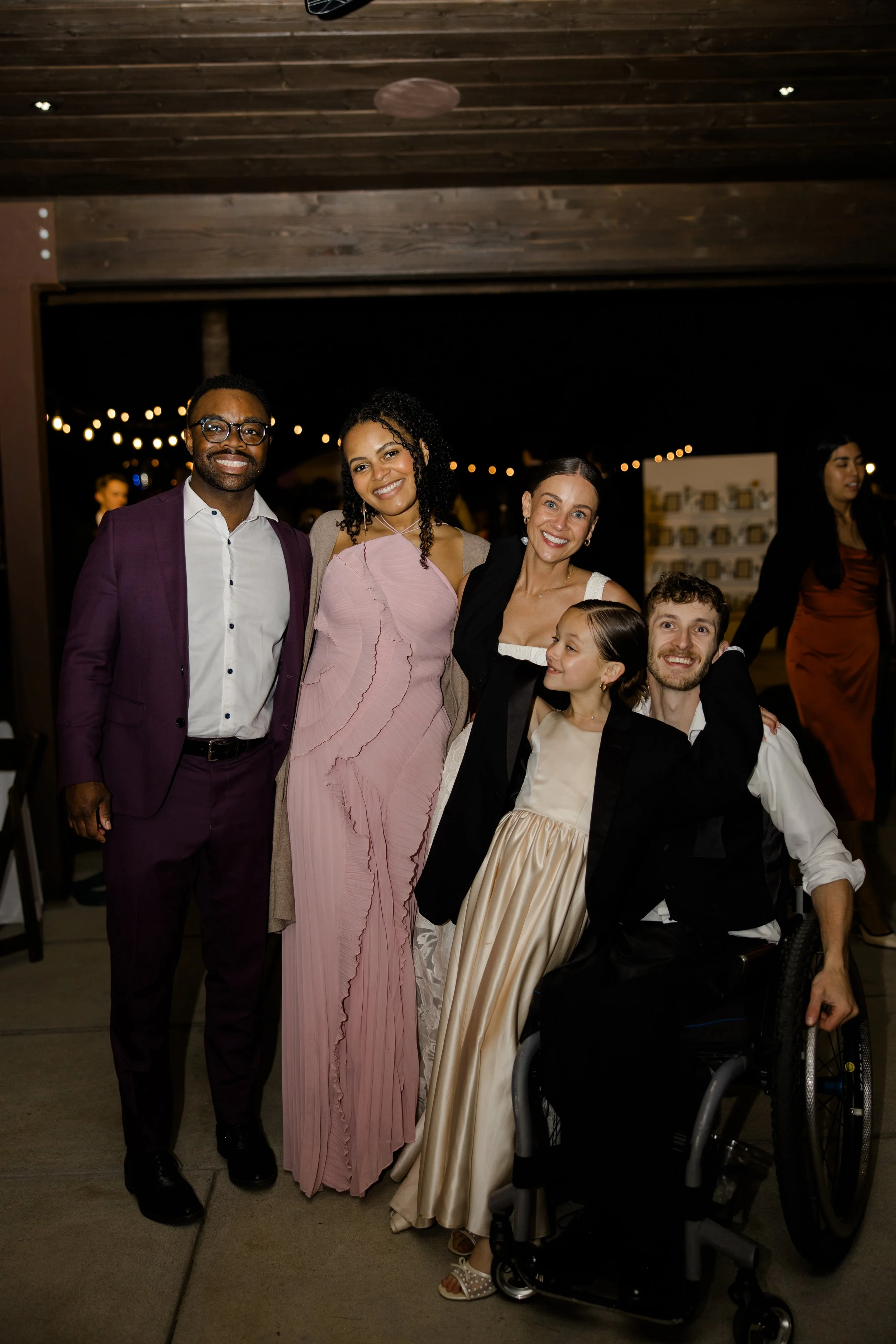 Group of five diverse individuals at an evening event, with outdoor string lights in the background. The group includes three women and two men, one man in a suit, and one woman in a wheelchair, all smiling and dressed in formal attire.