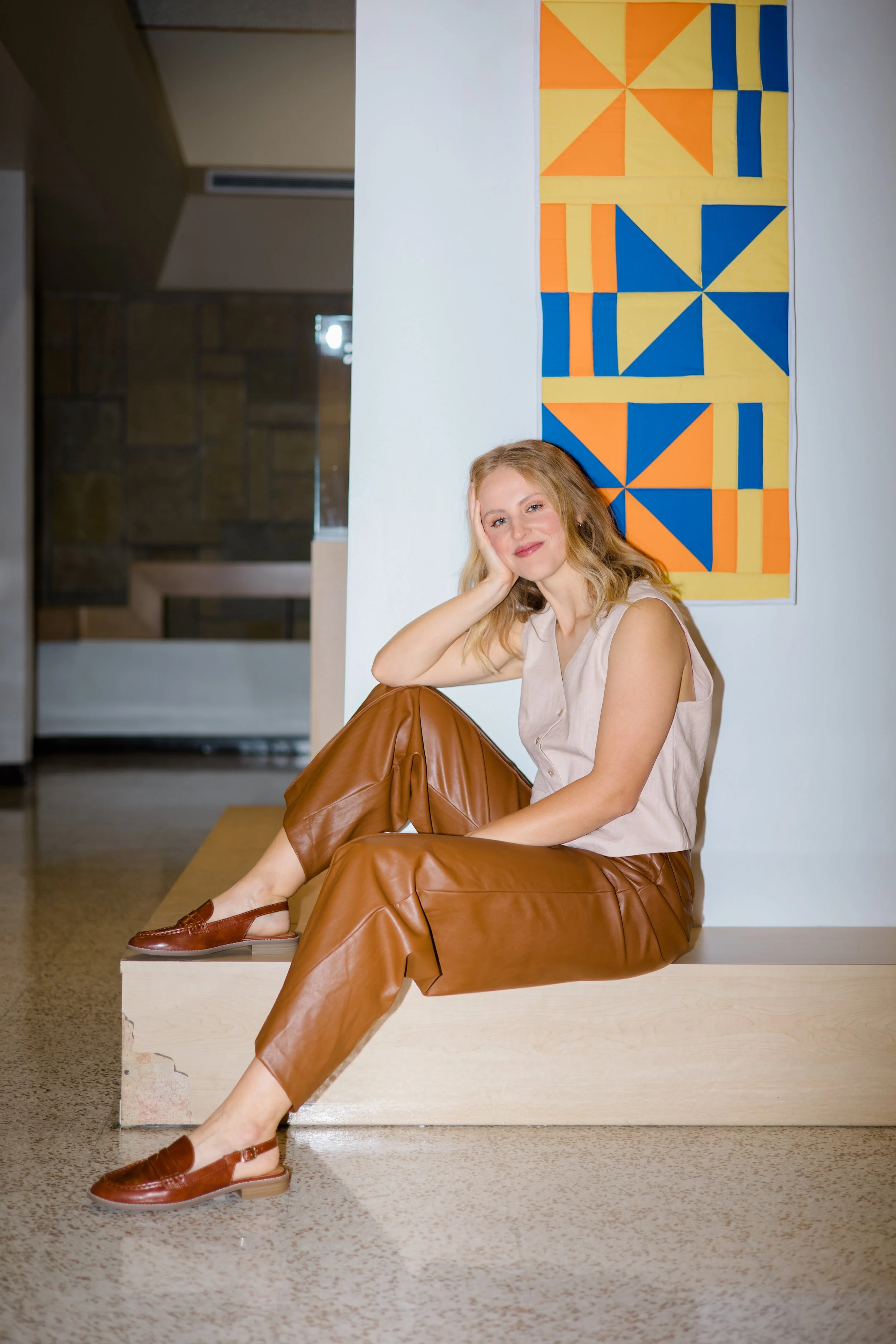 A woman with blonde hair sitting on a wooden block, smiling, with her hand supporting her head, wearing a beige sleeveless top, brown leather pants, and brown slip-on shoes. She is sitting in front of a geometric textile wall hanging with orange, yel