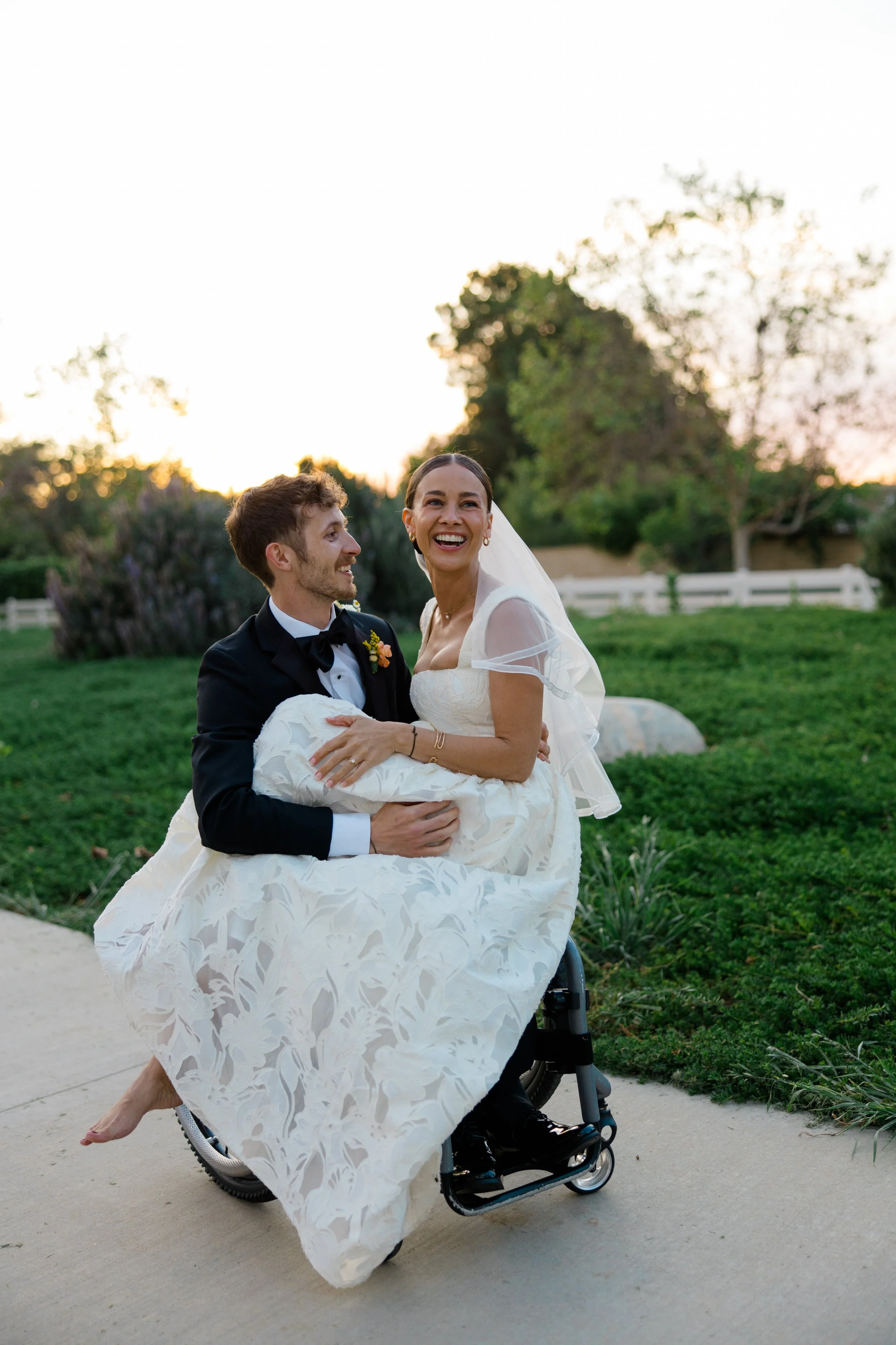 A bride in a wedding dress and smiling groom in a tuxedo are outdoors during sunset. The bride is sitting in a wheelchair, and the groom is holding her, lifting her slightly. They are surrounded by greenery, and a white fence and trees are in the bac