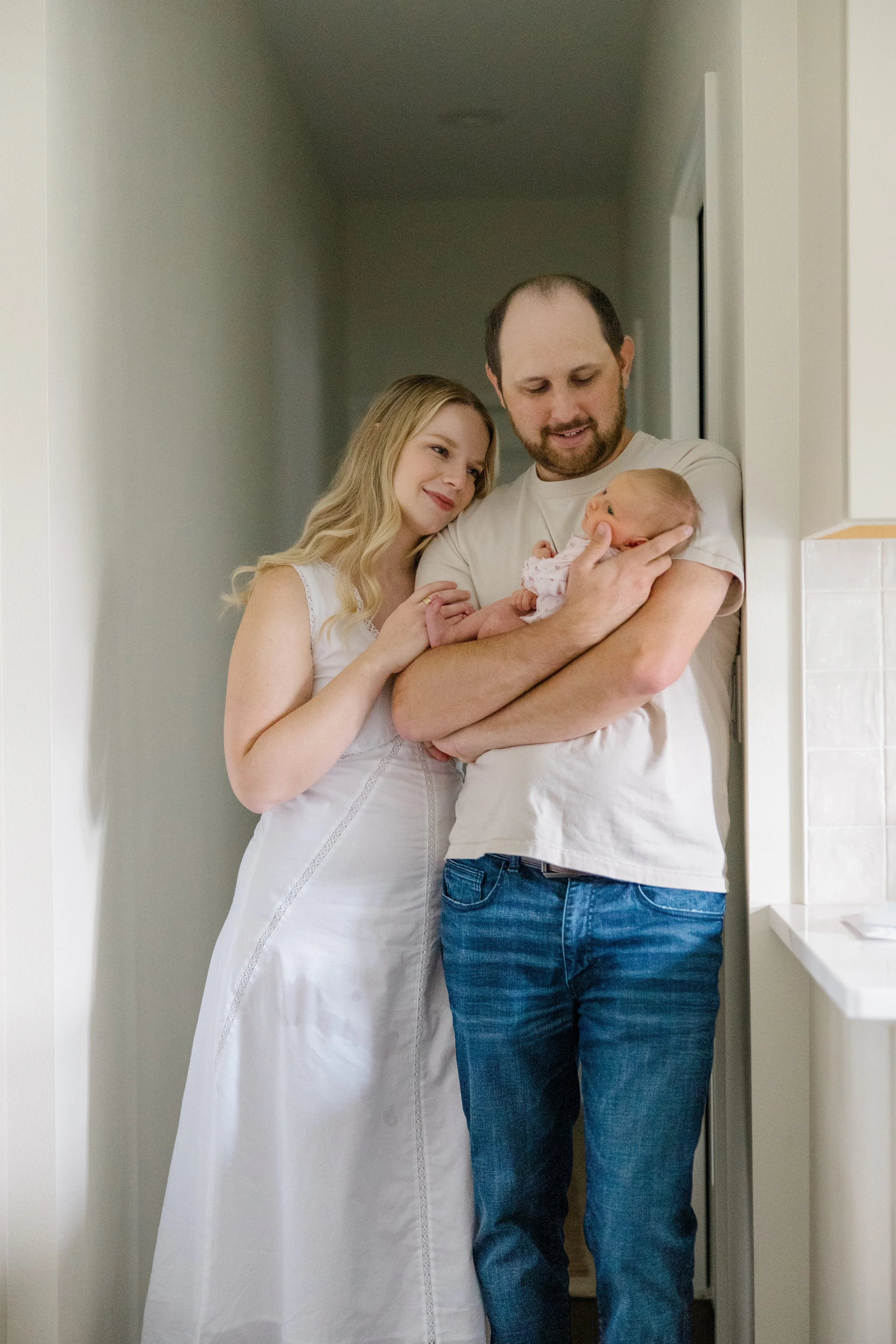 A woman in a white dress, a man in a white t-shirt and jeans, and a newborn baby in a pink patterned outfit standing in a hallway. The woman and man are looking at the baby lovingly.