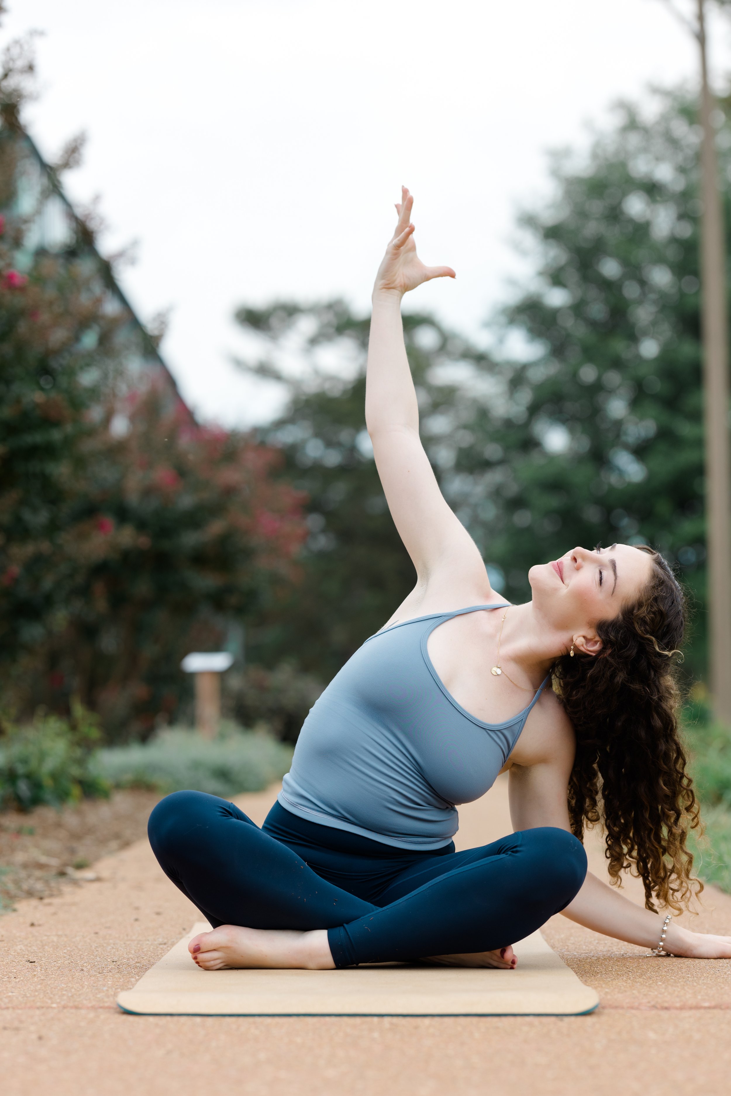 A woman practicing yoga outdoors, sitting cross-legged on a yoga mat, with her hands in a prayer position above her head, surrounded by trees and greenery.