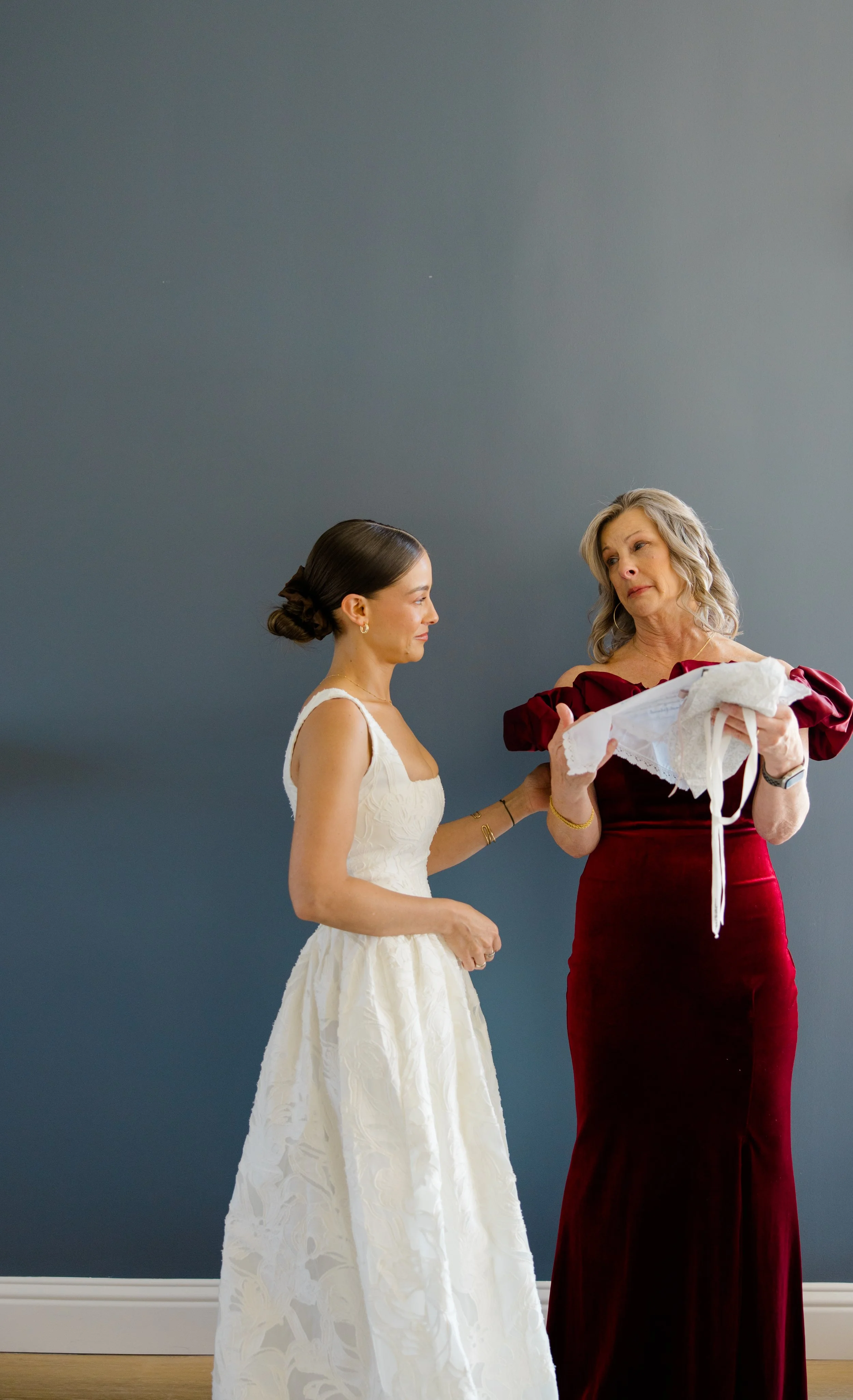 A woman in a white wedding dress stands next to another woman in a red velvet dress who is holding a lace wedding garter. They are in a room with a dark gray wall and wooden floor.