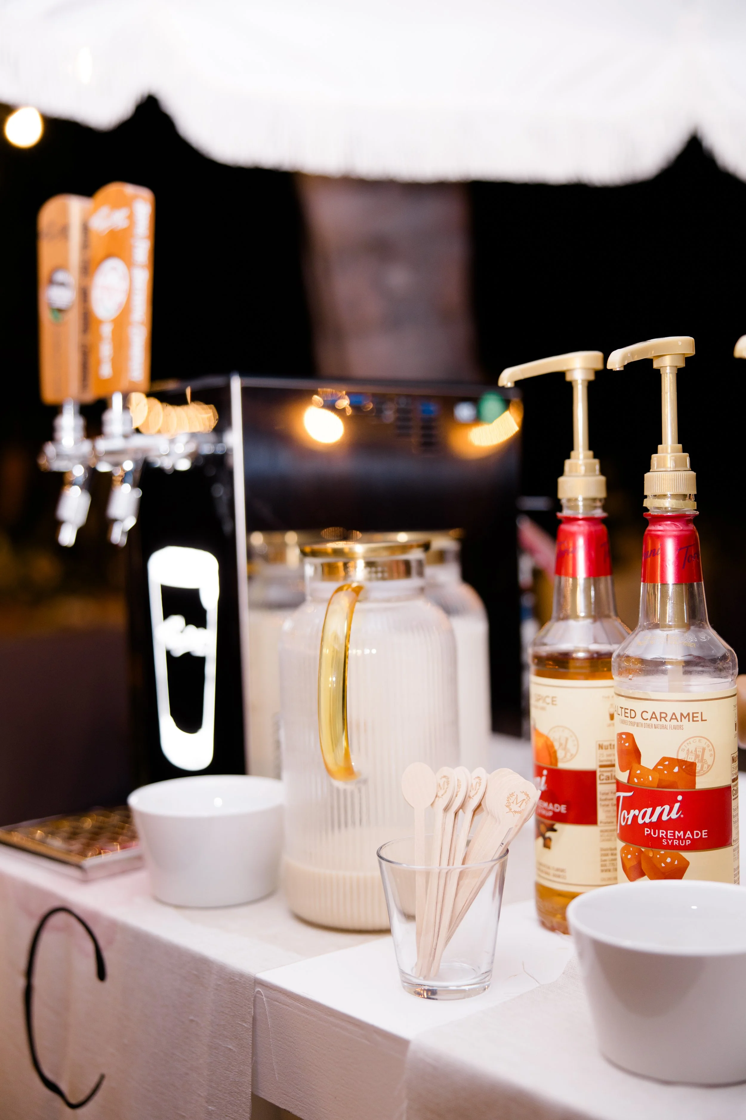 Close-up of a drinks station with syrup bottles, cups, a glass with a lemon slice, and various beverage dispensers set up on a table.