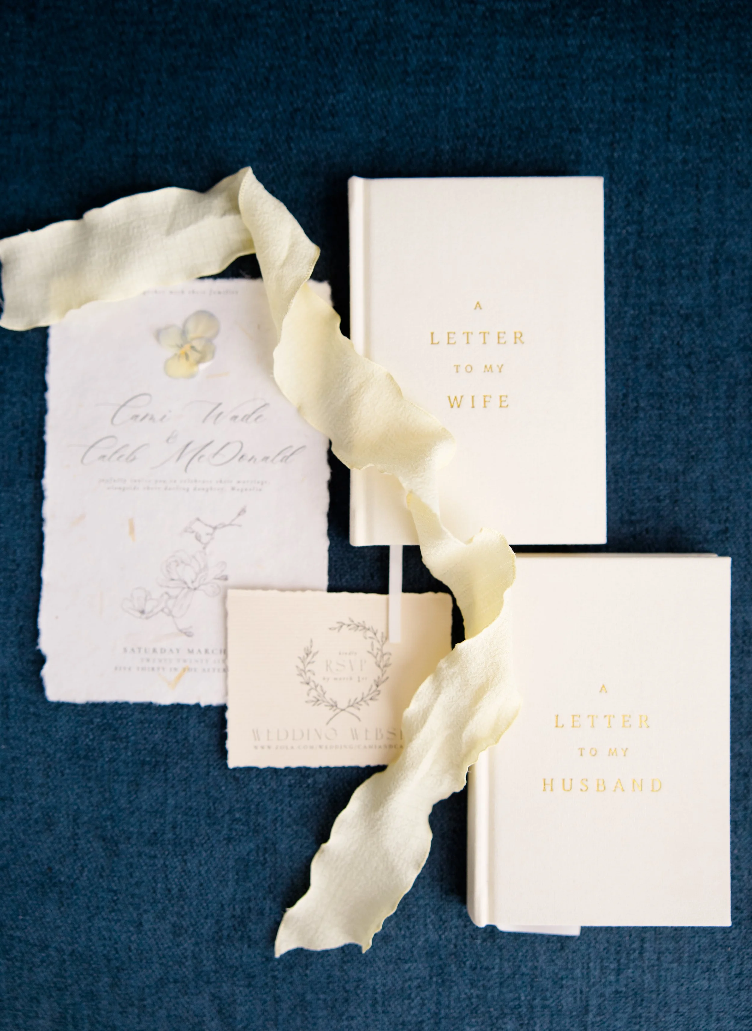 A white booklet titled 'A Letter to My Wife' with gold lettering, surrounded by wedding invitations and a torn piece of light yellow ribbon on a dark blue fabric background.