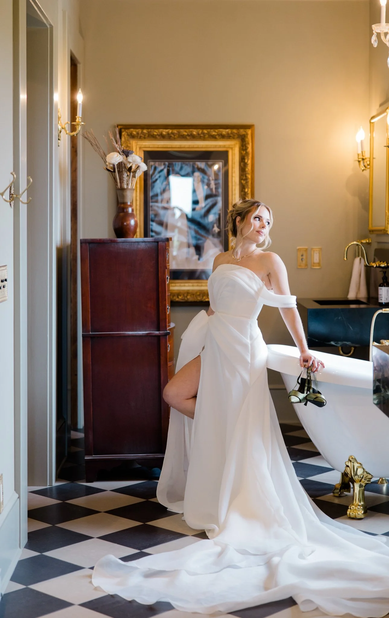 A woman in a white wedding dress standing in a room with a black and white checkered floor. She is leaning on a bathtub, holding a pair of shoes, and looking away. The room has gold-framed mirrors, a vase with dried flowers, and elegant wall sconces.