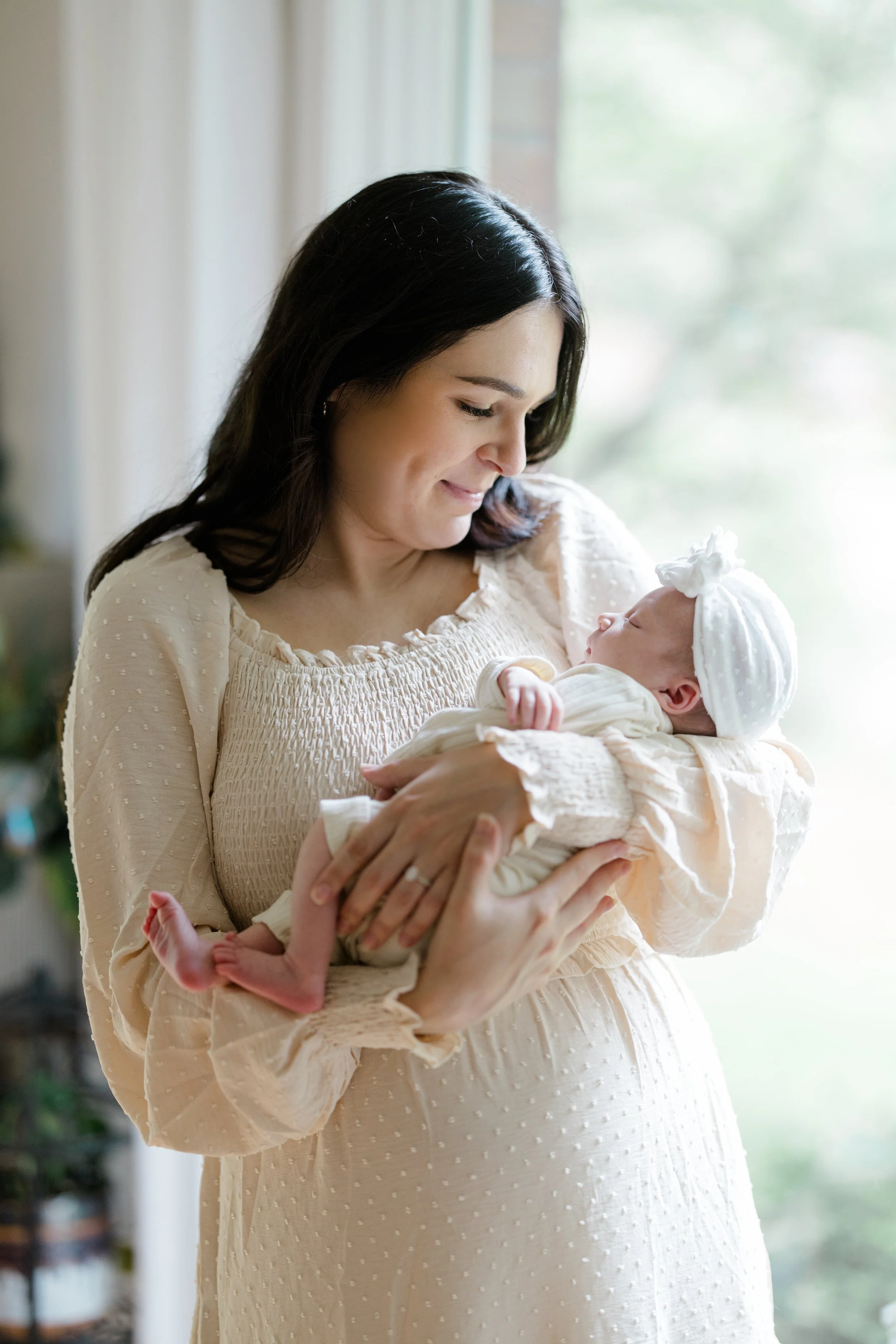 A woman with dark hair in a light beige, textured dress holding a newborn baby wrapped in a matching blanket near a window with a soft smile, looking down at the baby.
