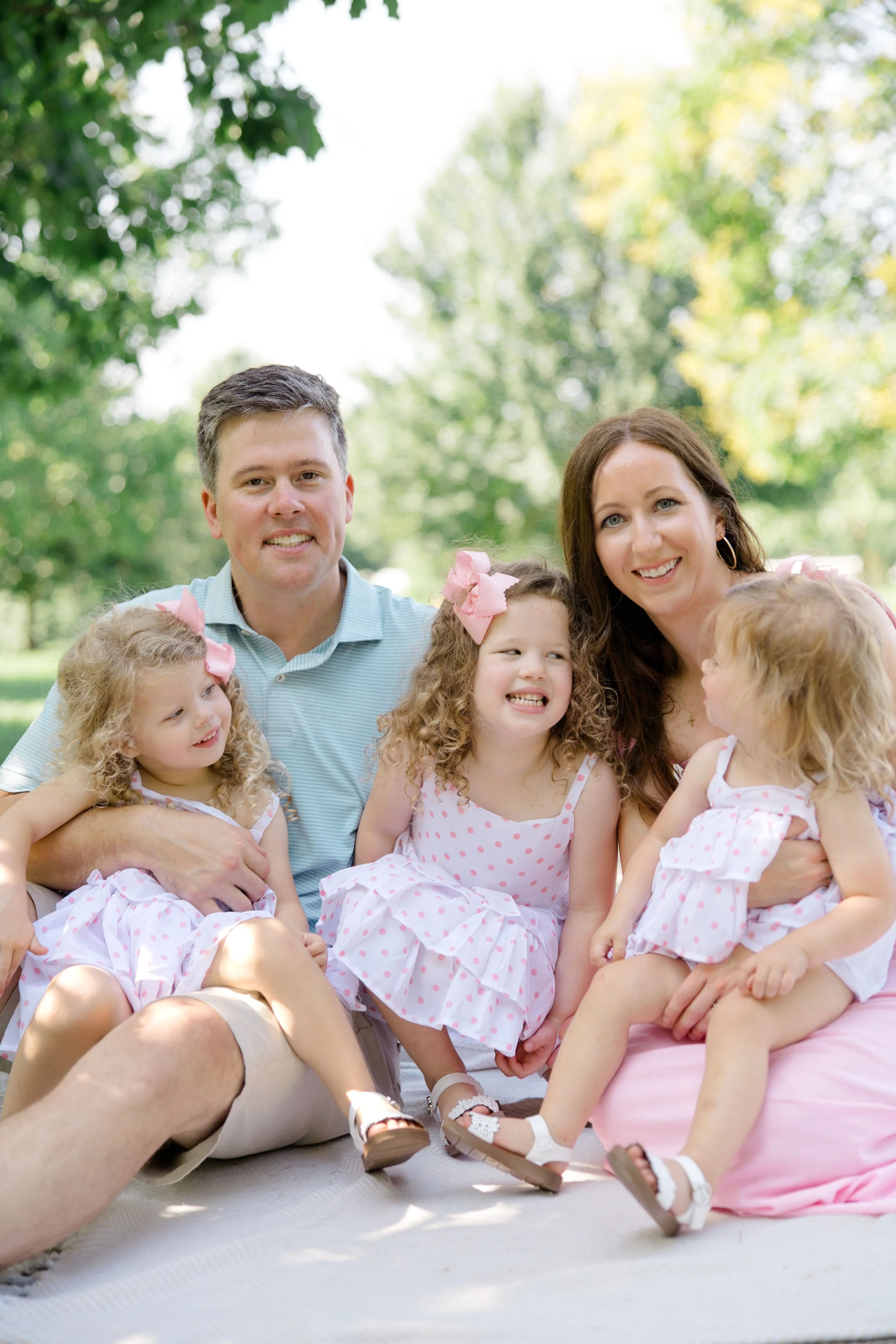 A family of six sitting outdoors on a sunny day. The group includes two adults and four young children, all dressed in light, summery clothing with pink accents. They are smiling and appear to be enjoying each other's company against a background of 