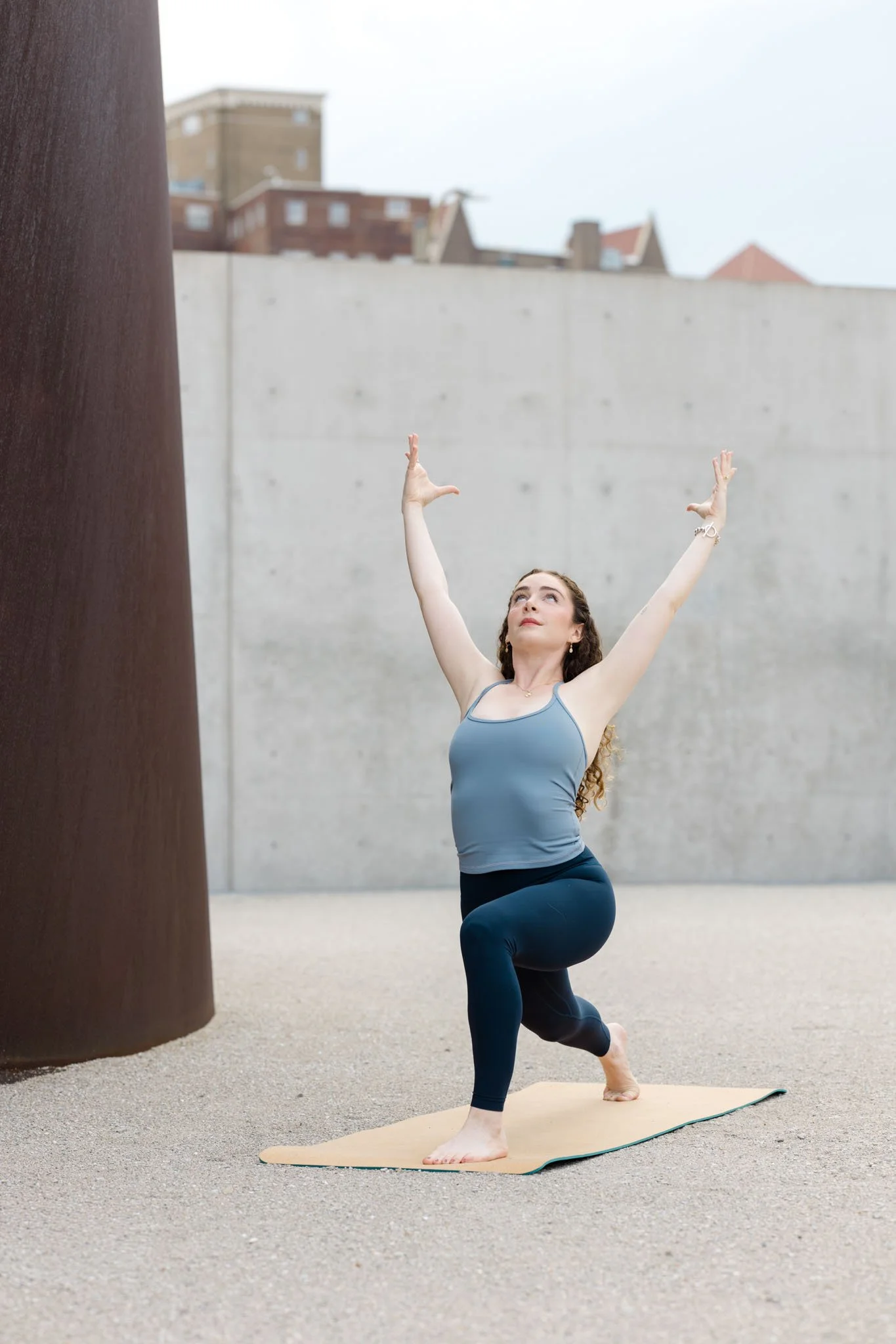 A woman practicing yoga outdoors on a beige yoga mat, performing a low lunge pose with her arms raised, near a large dark-colored sculpture with a cityscape in the background.