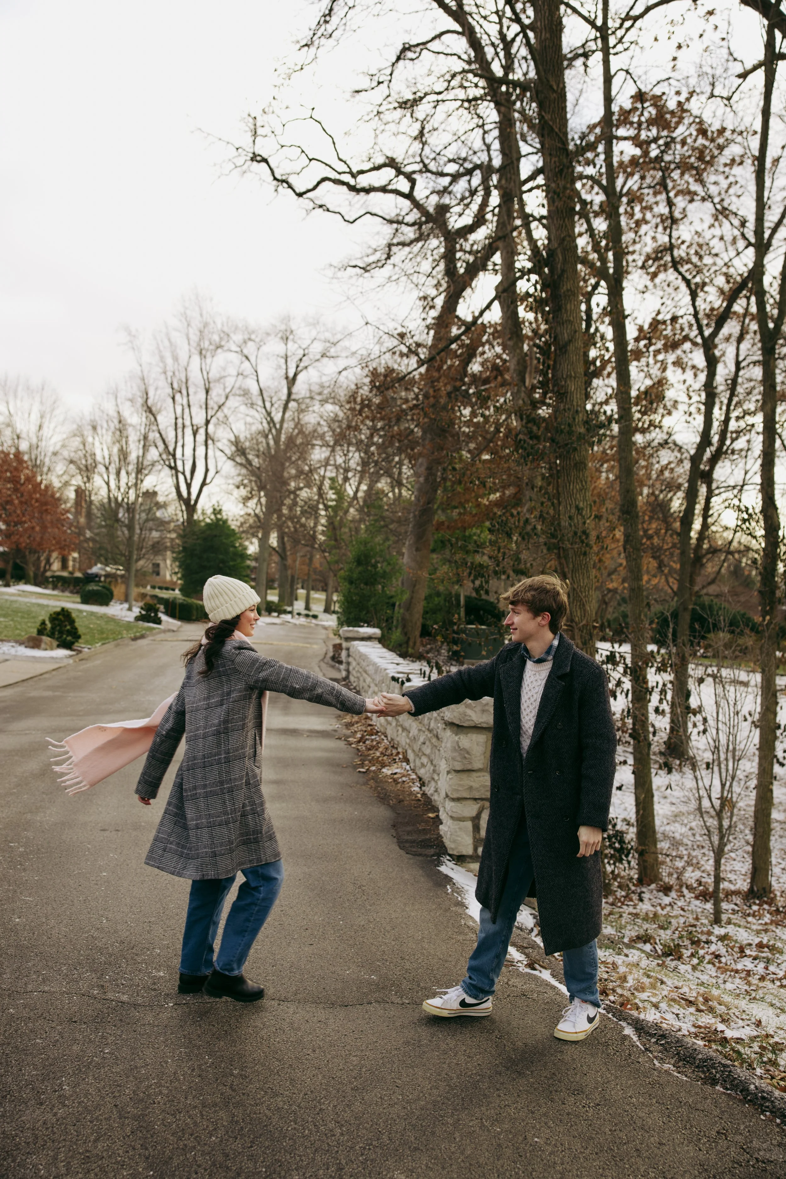 A young couple holding hands and dancing outdoors on a cloudy day in a park or residential area with bare trees and patches of snow on the ground.