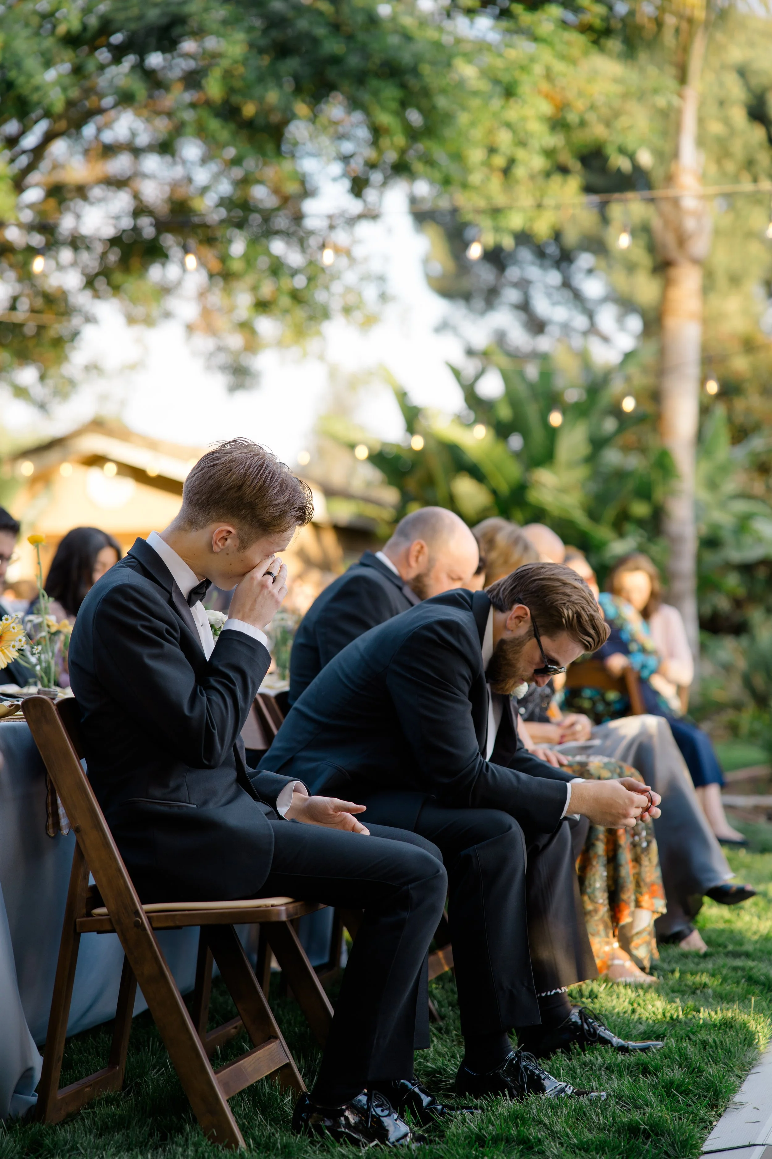 People seated outdoors at a wedding, wearing formal attire, some with heads bowed and eyes closed in prayer.