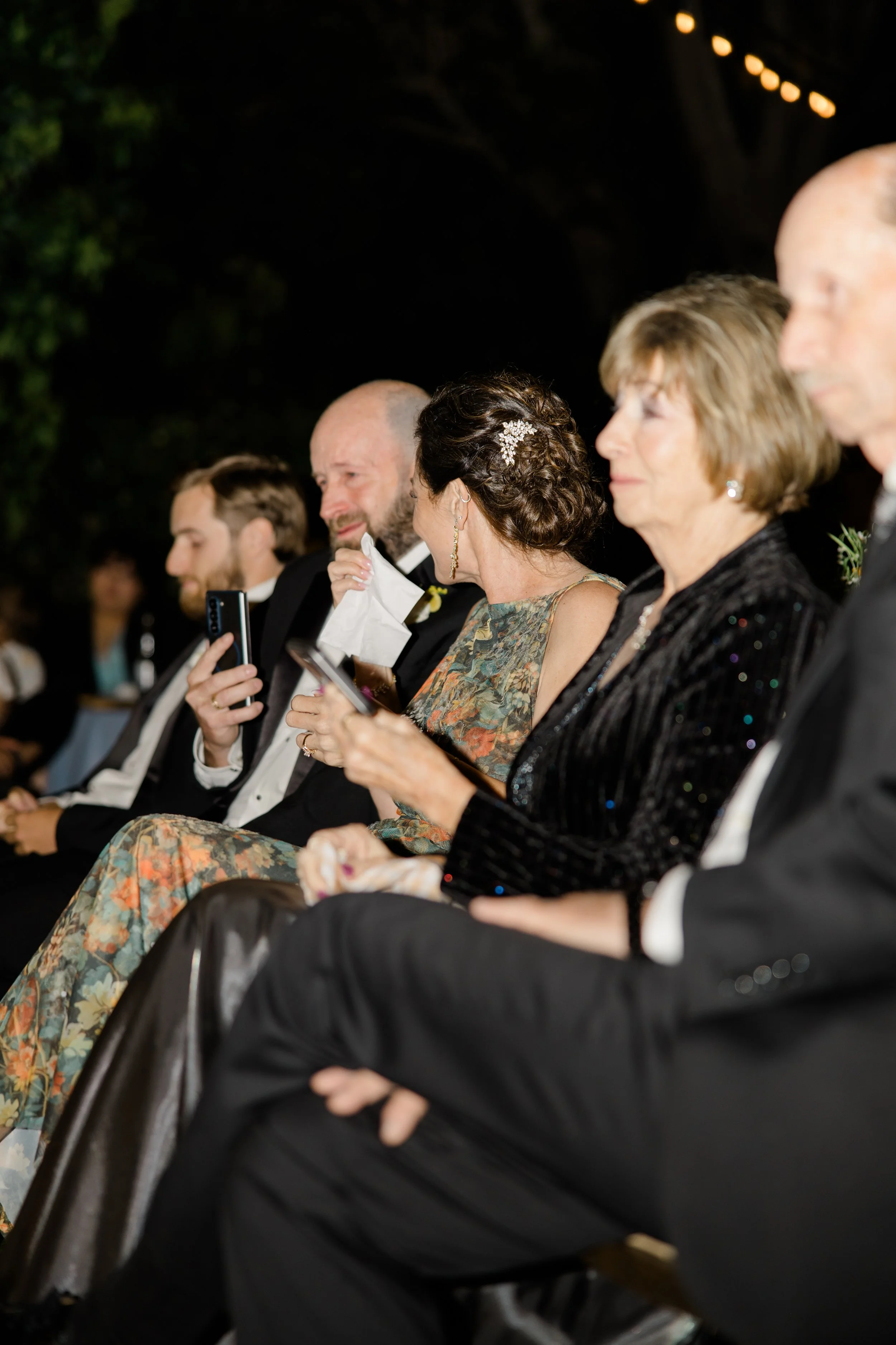 People seated at an outdoor event in the evening, some using their phones, with dark background and string lights overhead.