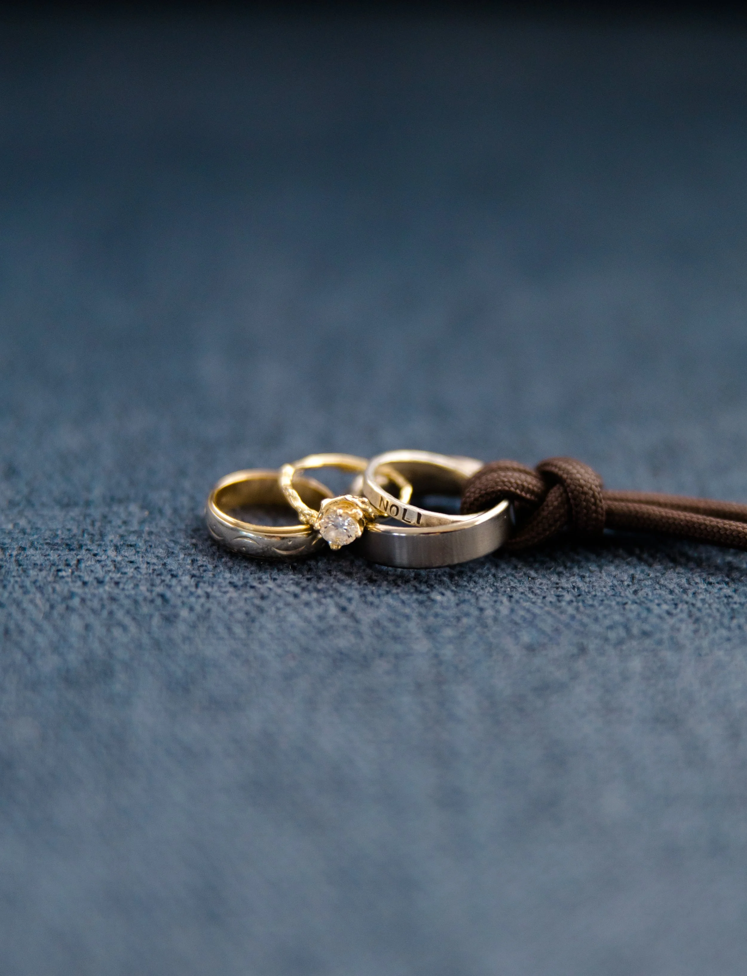 Set of rings, including an engagement ring with a diamond and a wedding band, placed on a dark fabric background with a cord nearby.