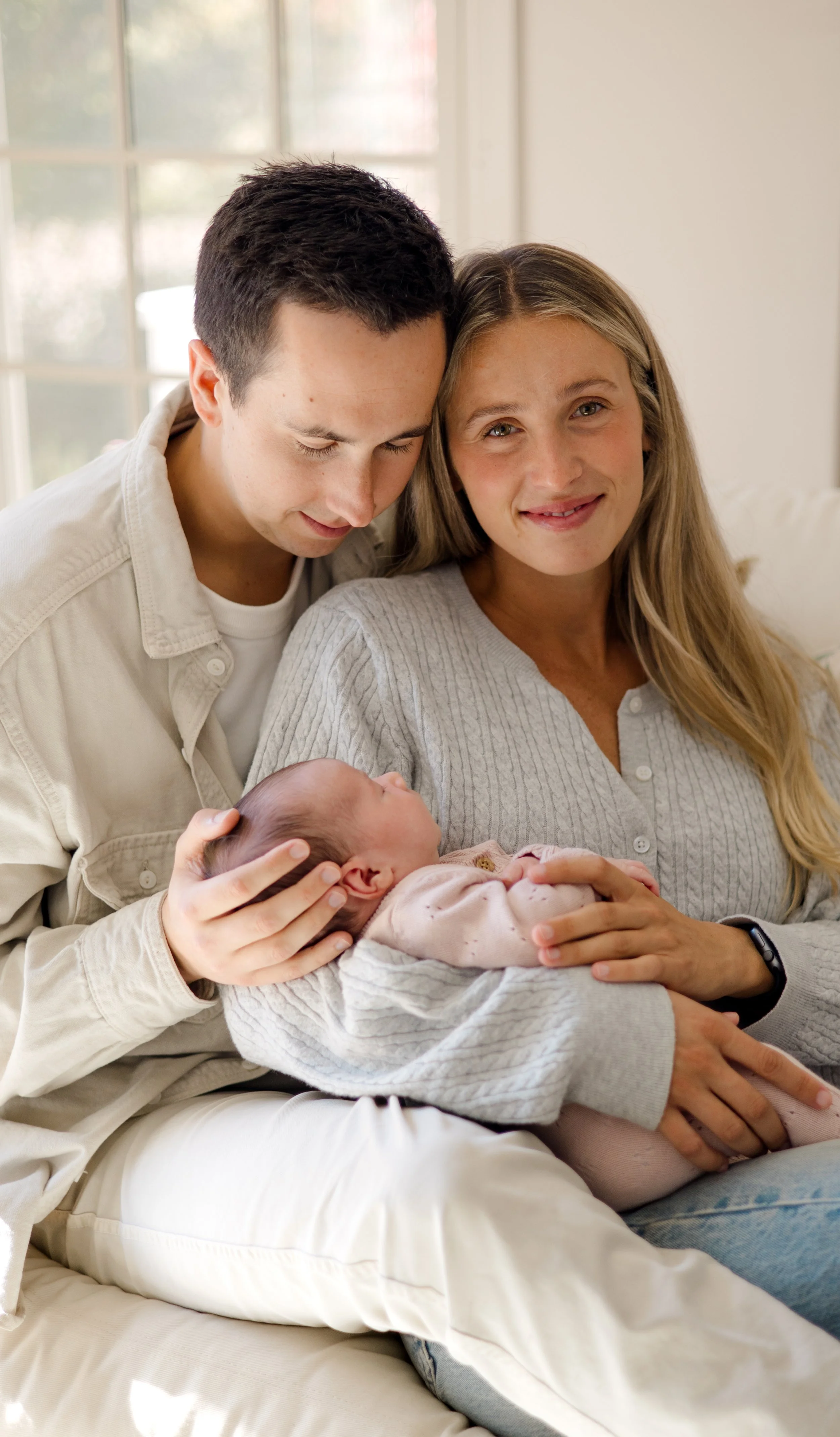 A family with a young woman, a young man, and a newborn baby in a cozy, sunlit room. The young woman is holding the baby, smiling at the camera while the young man gently rests his head against her and looks down at the baby.