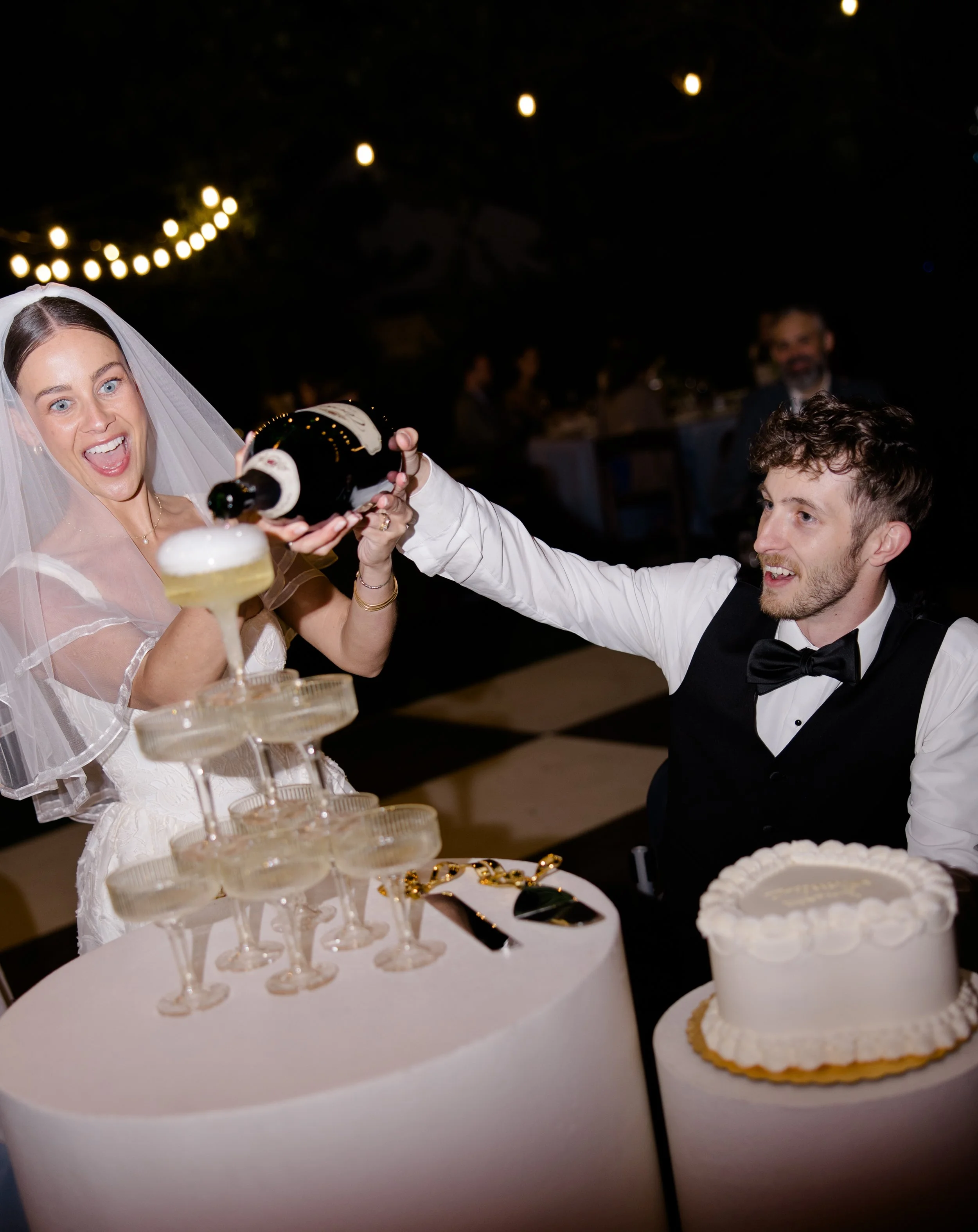 A bride in a wedding dress and veil is smiling and holding a champagne bottle, pouring it into a glass held by a groom in a tuxedo at a wedding reception. There is a table with a multi-tiered cake and glasses of champagne.
