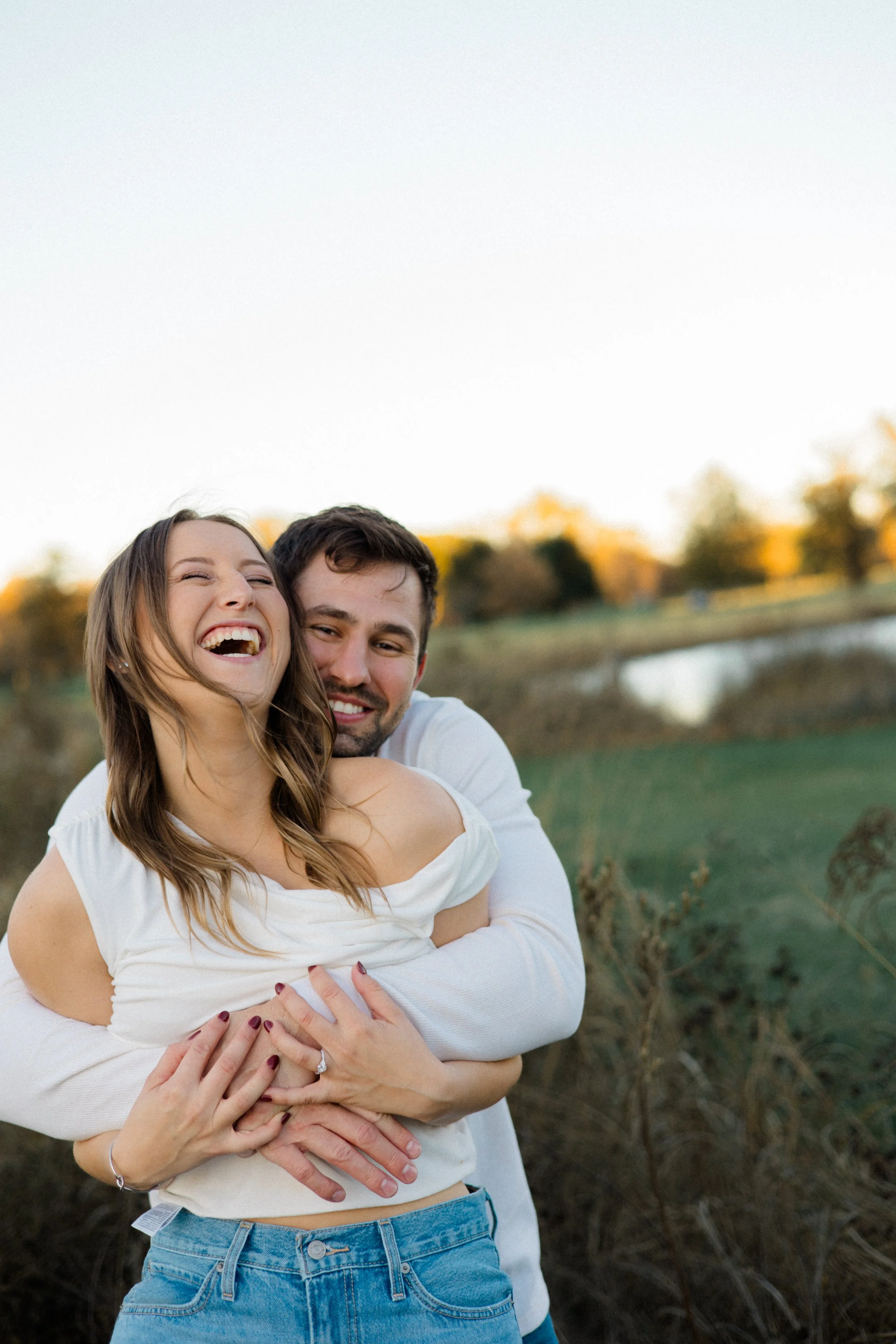A happy couple laughing and embracing outdoors near a pond at sunset with trees in the background.