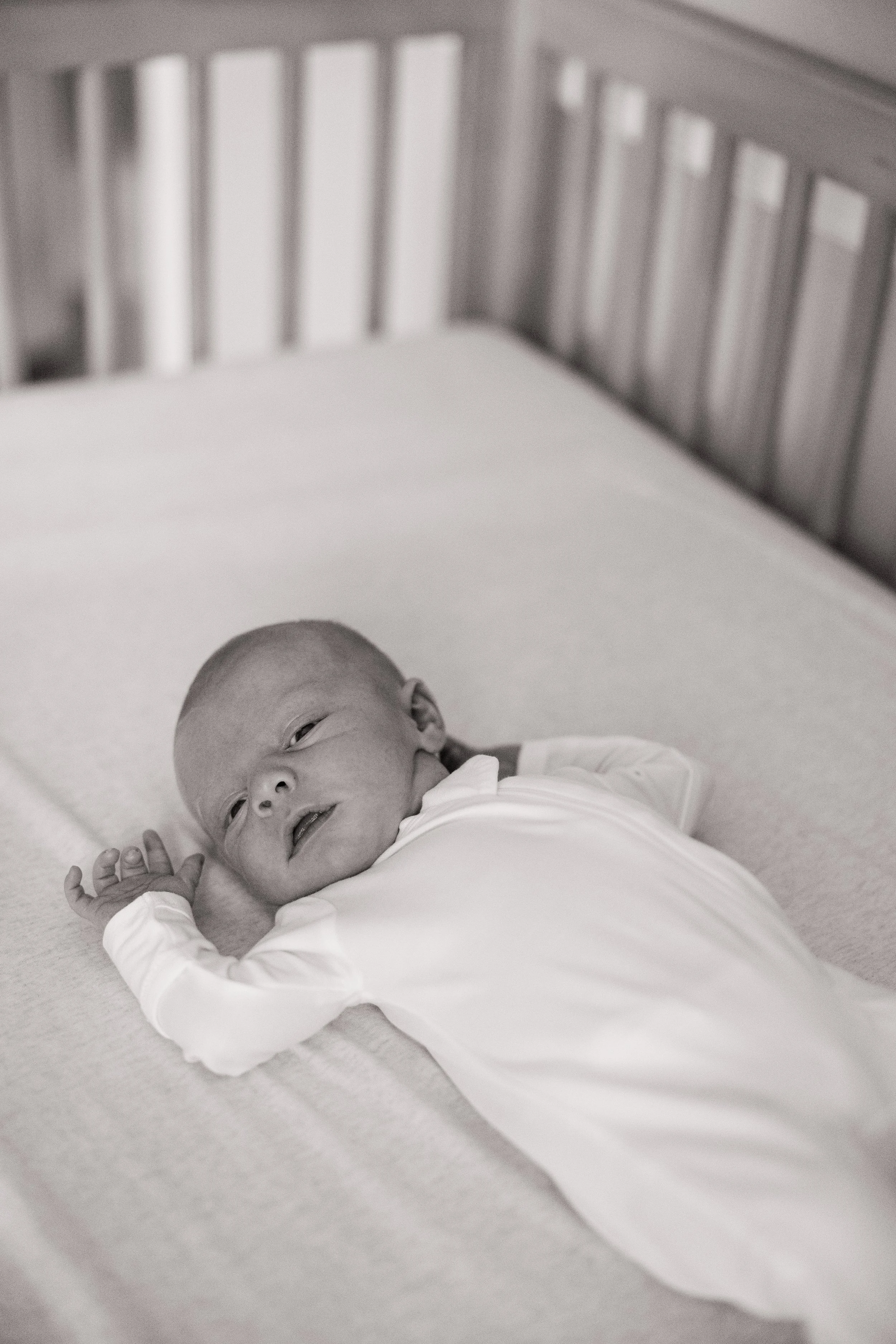 Black and white photo of a newborn baby lying on their back on a soft surface, looking at the camera with their left hand near their face, and a crib in the background.