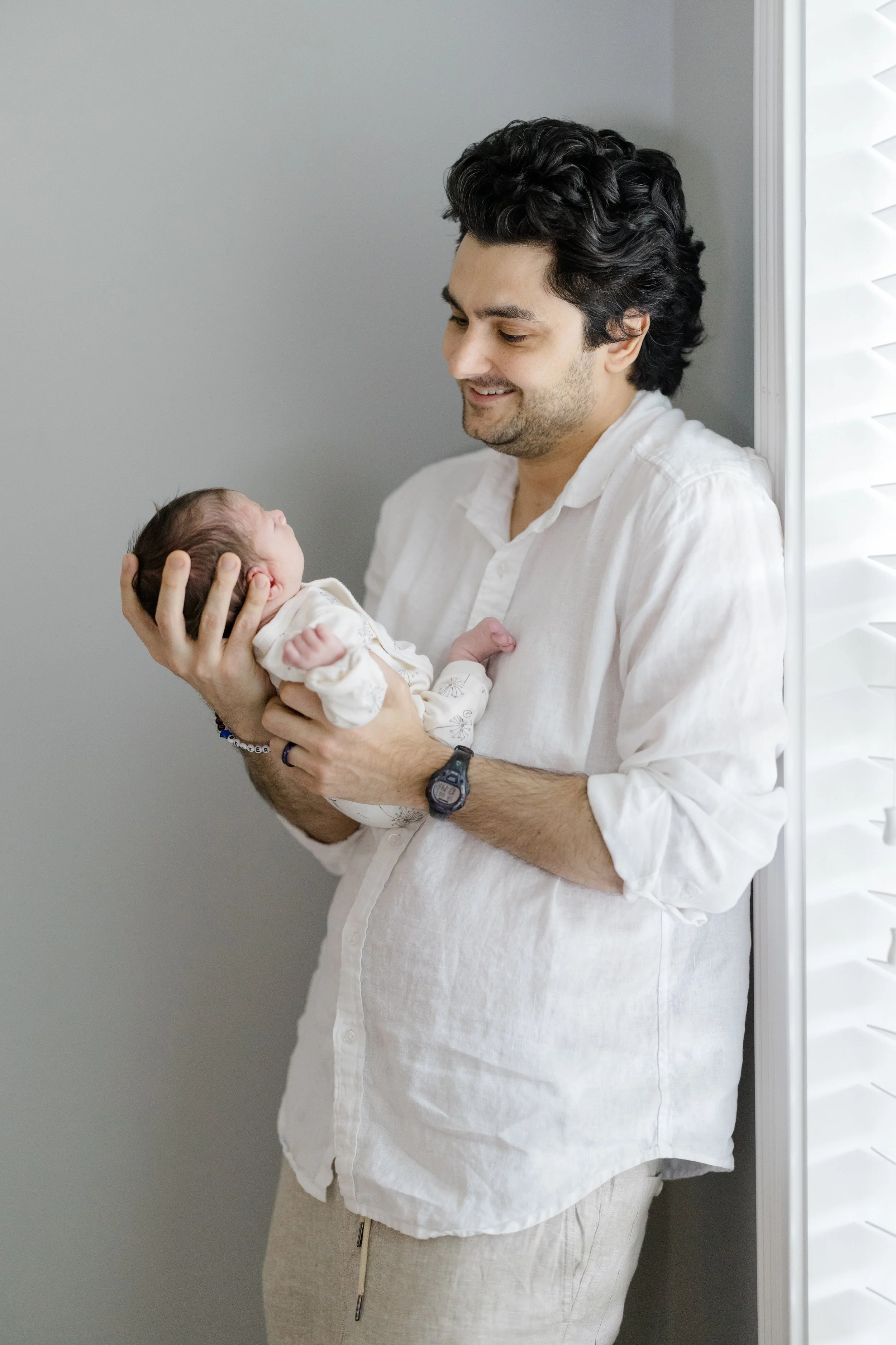 A man with dark, curly hair wearing a white shirt holds a newborn baby in his hands while standing near a wall and a window with white blinds.