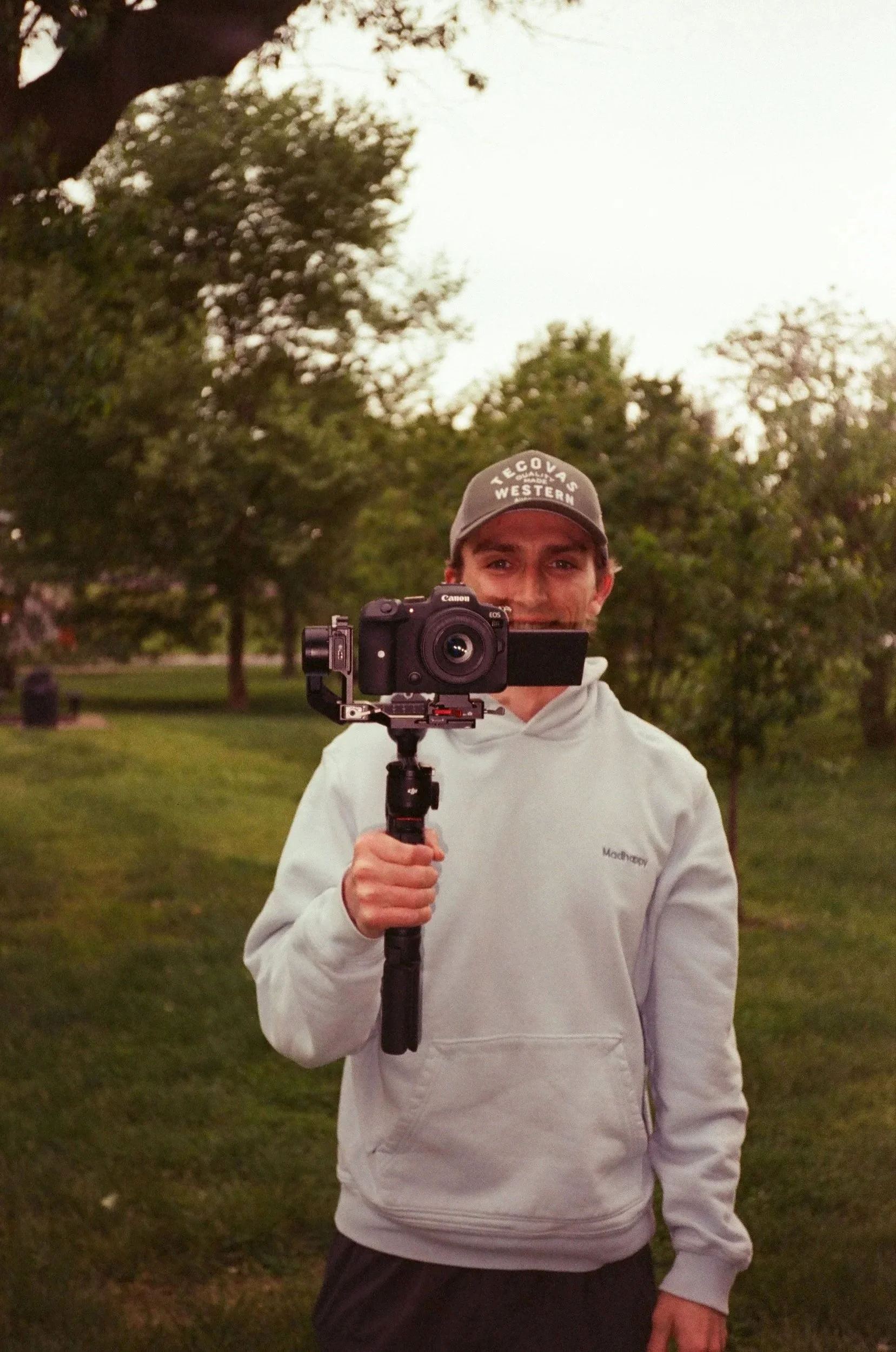 A young man wearing a gray cap with the words 'TECOS WESTERN' on it, and a white hoodie, is holding a camera stabilizer with a camera mounted on it, pointing towards the mirror in a park. The background features green trees and a grassy area, with a soft evening light.
