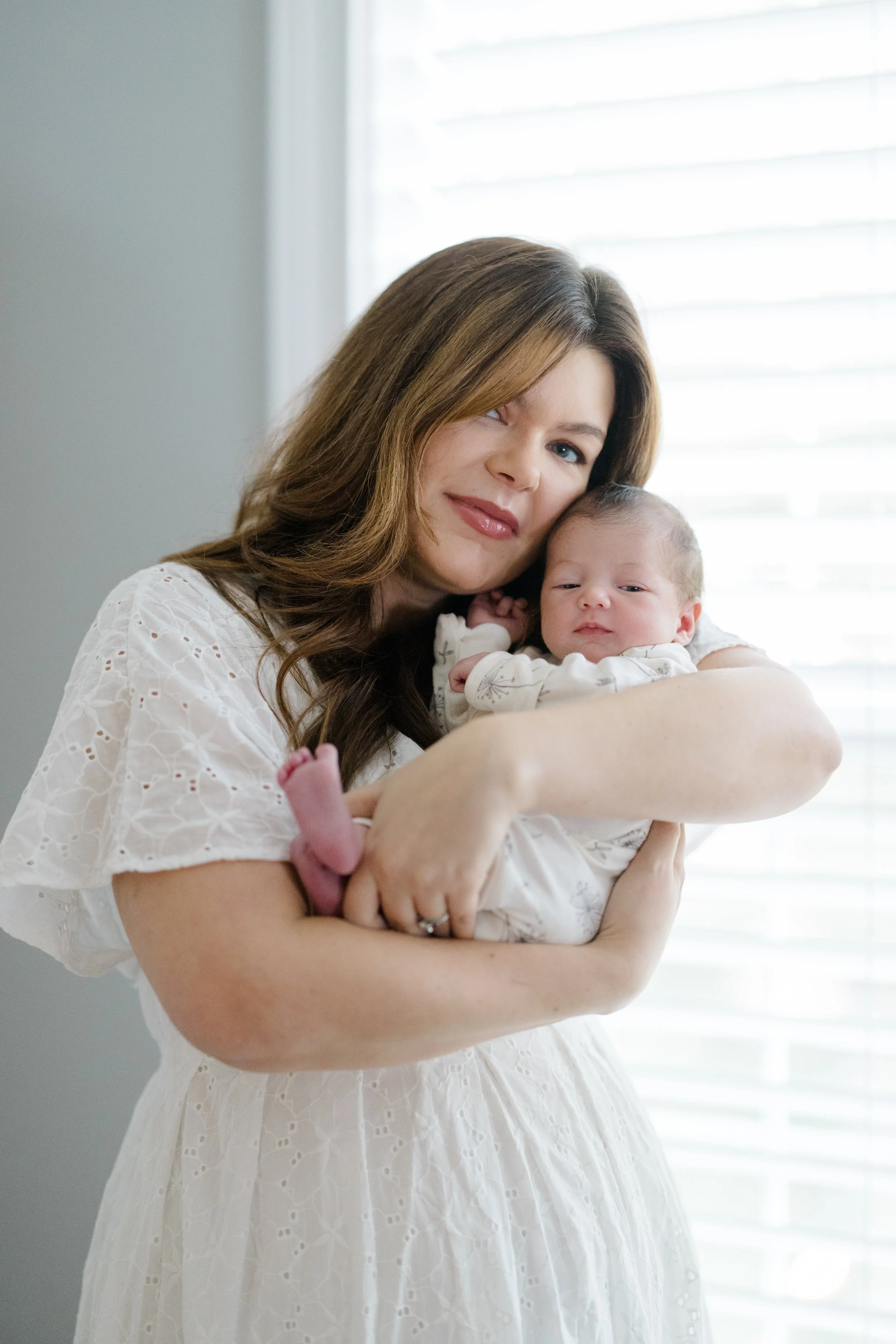 A woman with long brown hair holds a newborn baby in her arms, standing near a window with blinds that let in natural light.