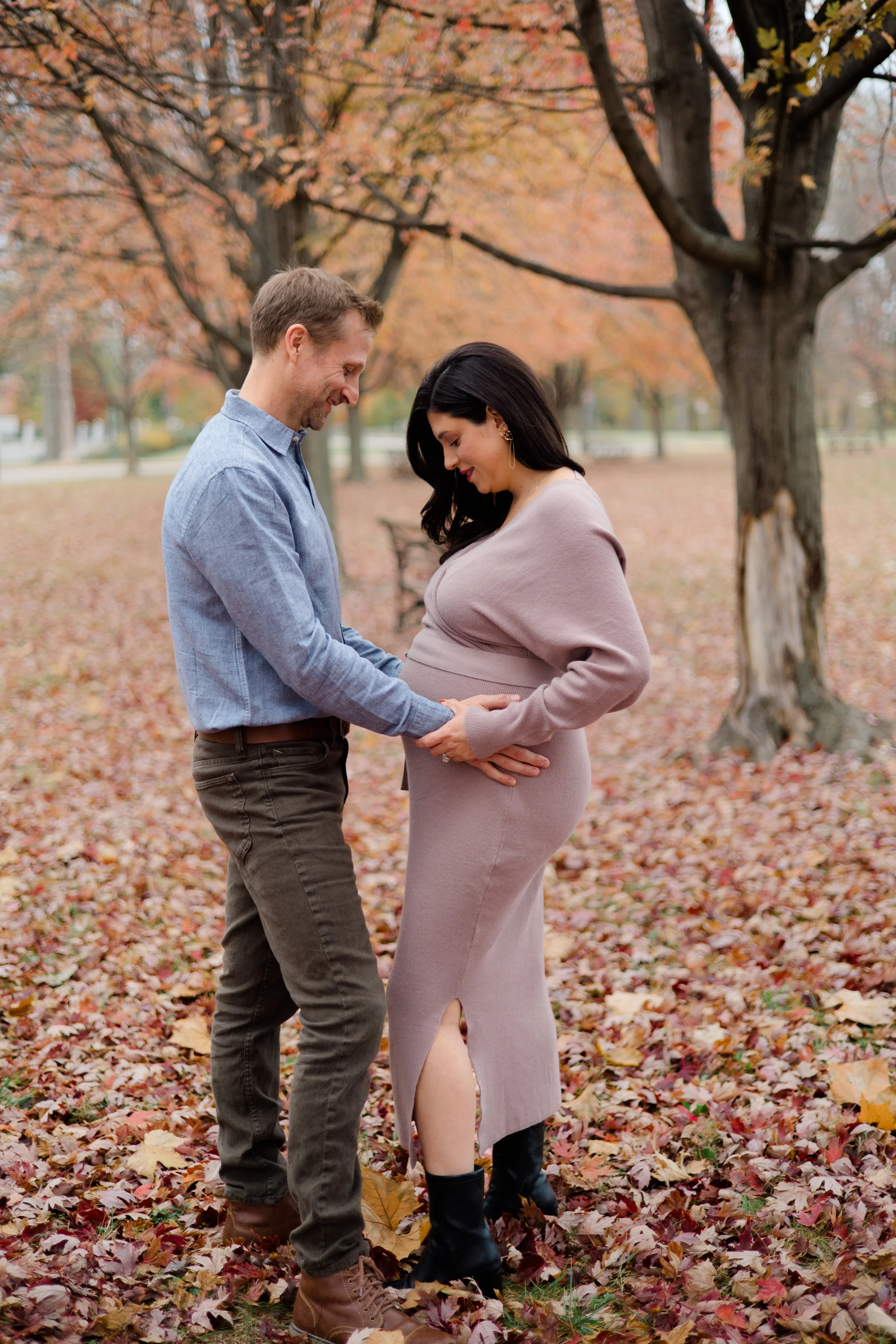 A couple standing in a park covered in fallen autumn leaves, gazing at each other lovingly with trees in the background.