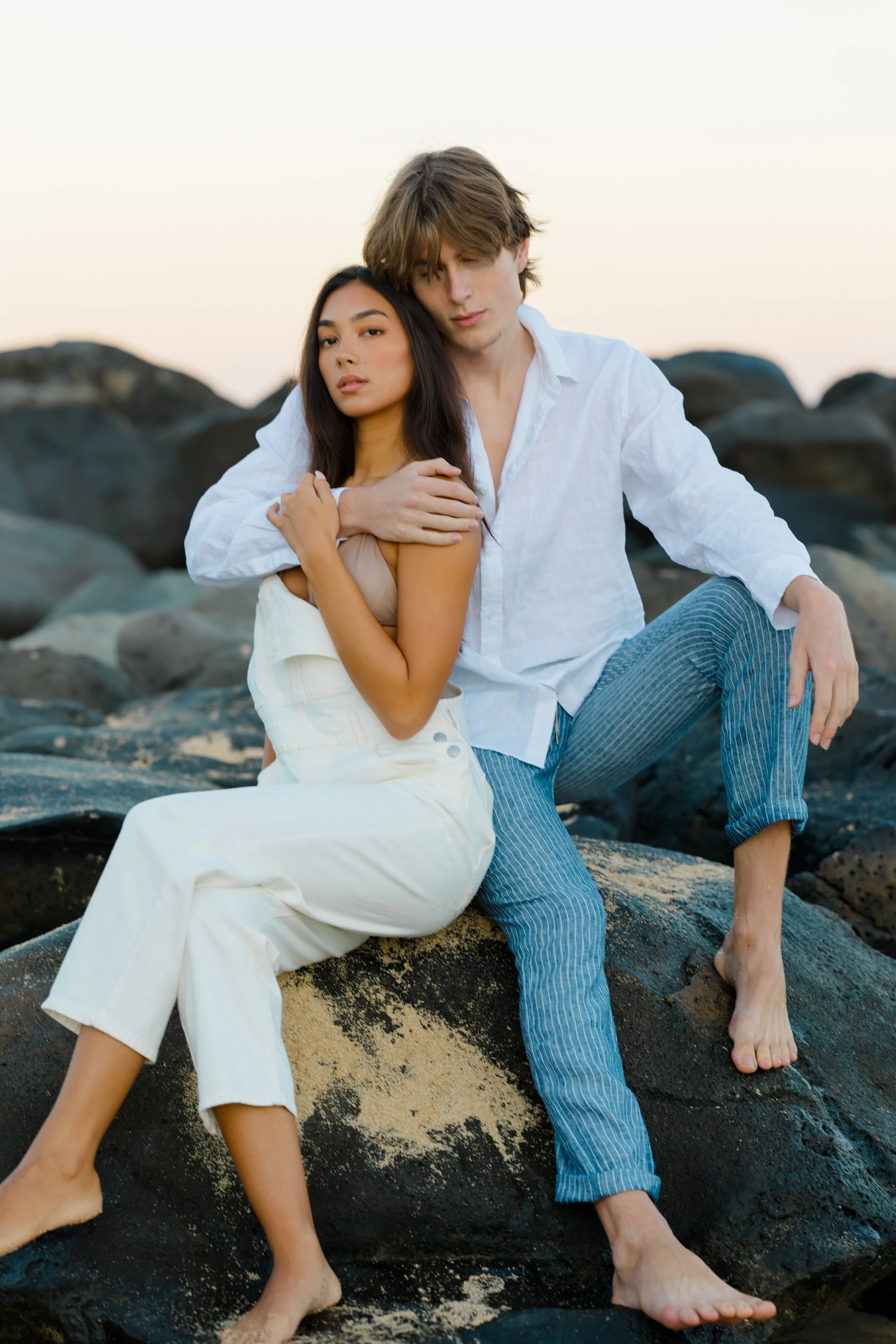 A young man and woman sitting on rocks by the beach at sunset, with the woman leaning against the man, both barefoot and dressed in casual white and light-colored clothing.