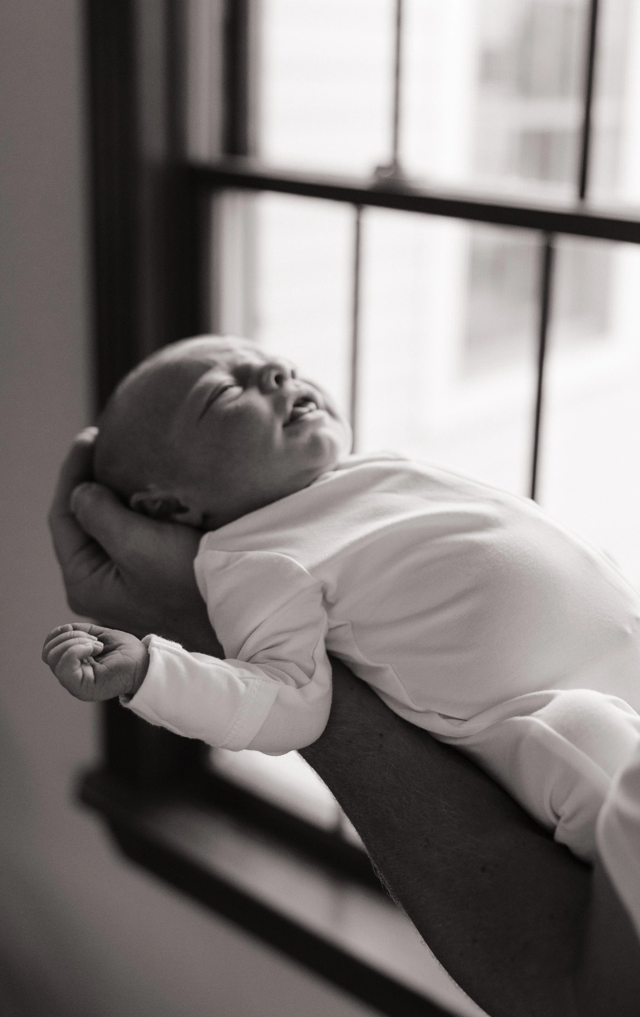 A baby dressed in white lying on a person's arm near a window.