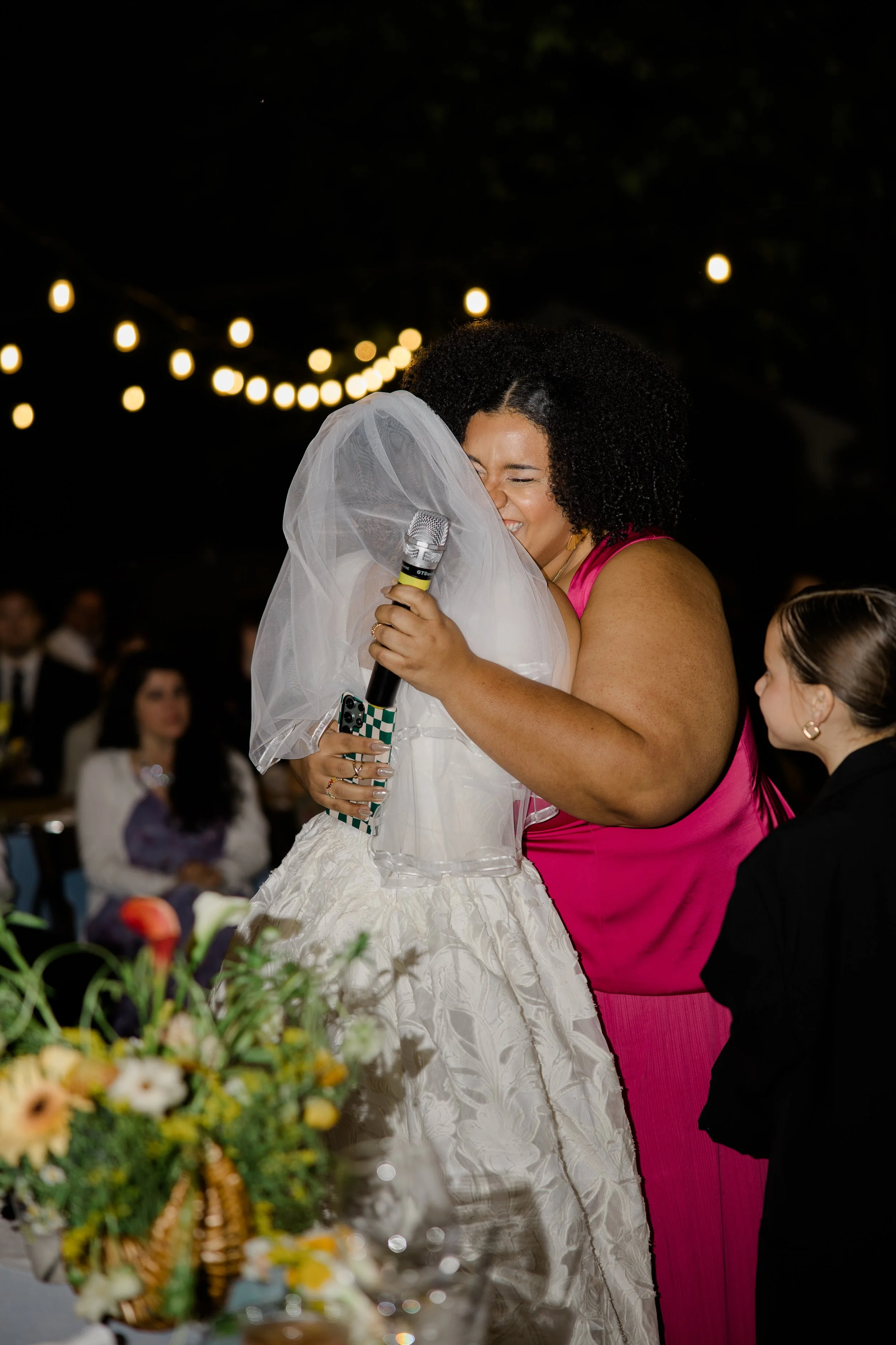 A woman in a pink dress embraces a bride in a white wedding gown and veil, holding a microphone at a nighttime outdoor wedding reception with string lights overhead.
