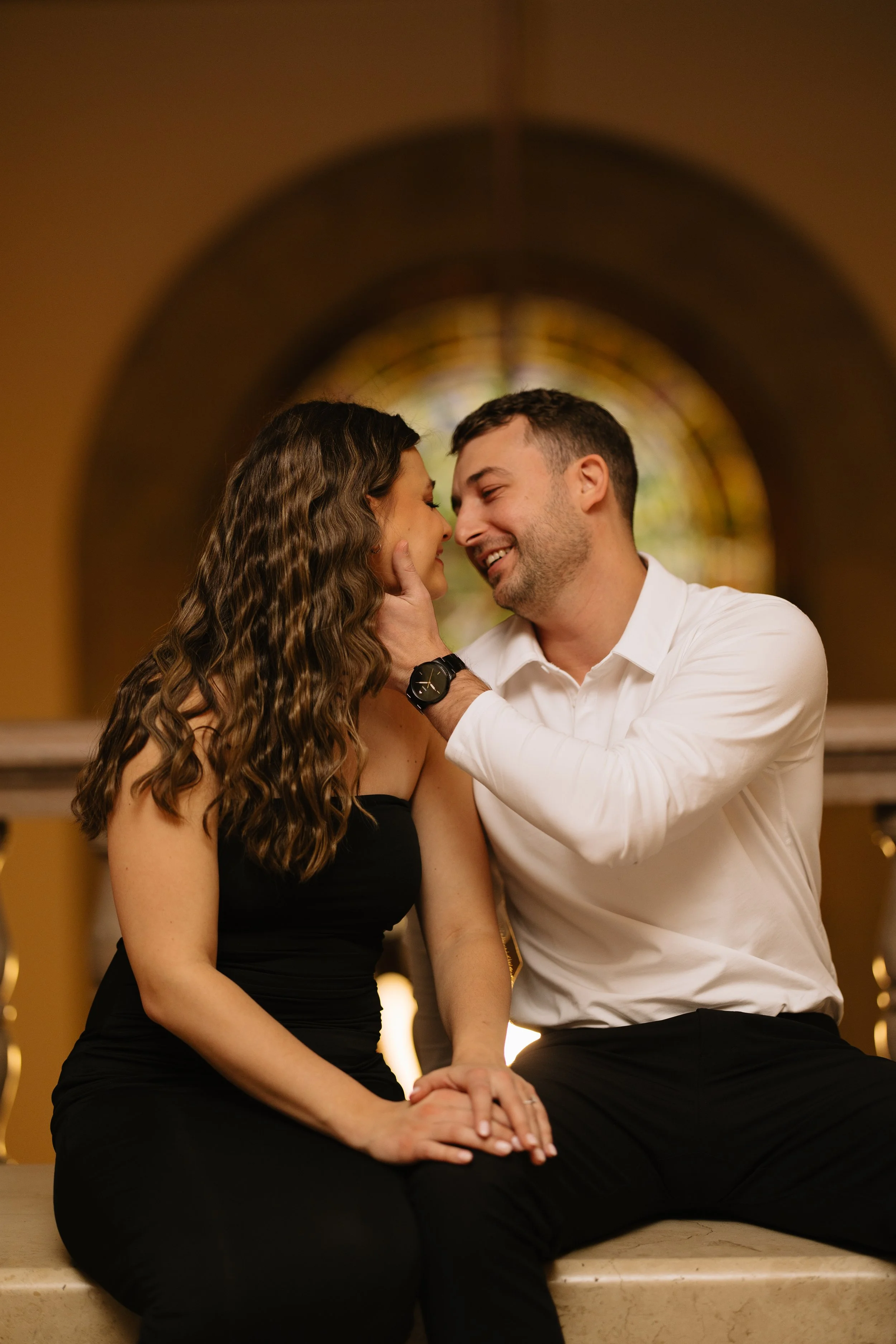 A couple sitting on a bench in an artsy setting, with the man touching the woman's face gently as they smile at each other, behind a stained glass window.