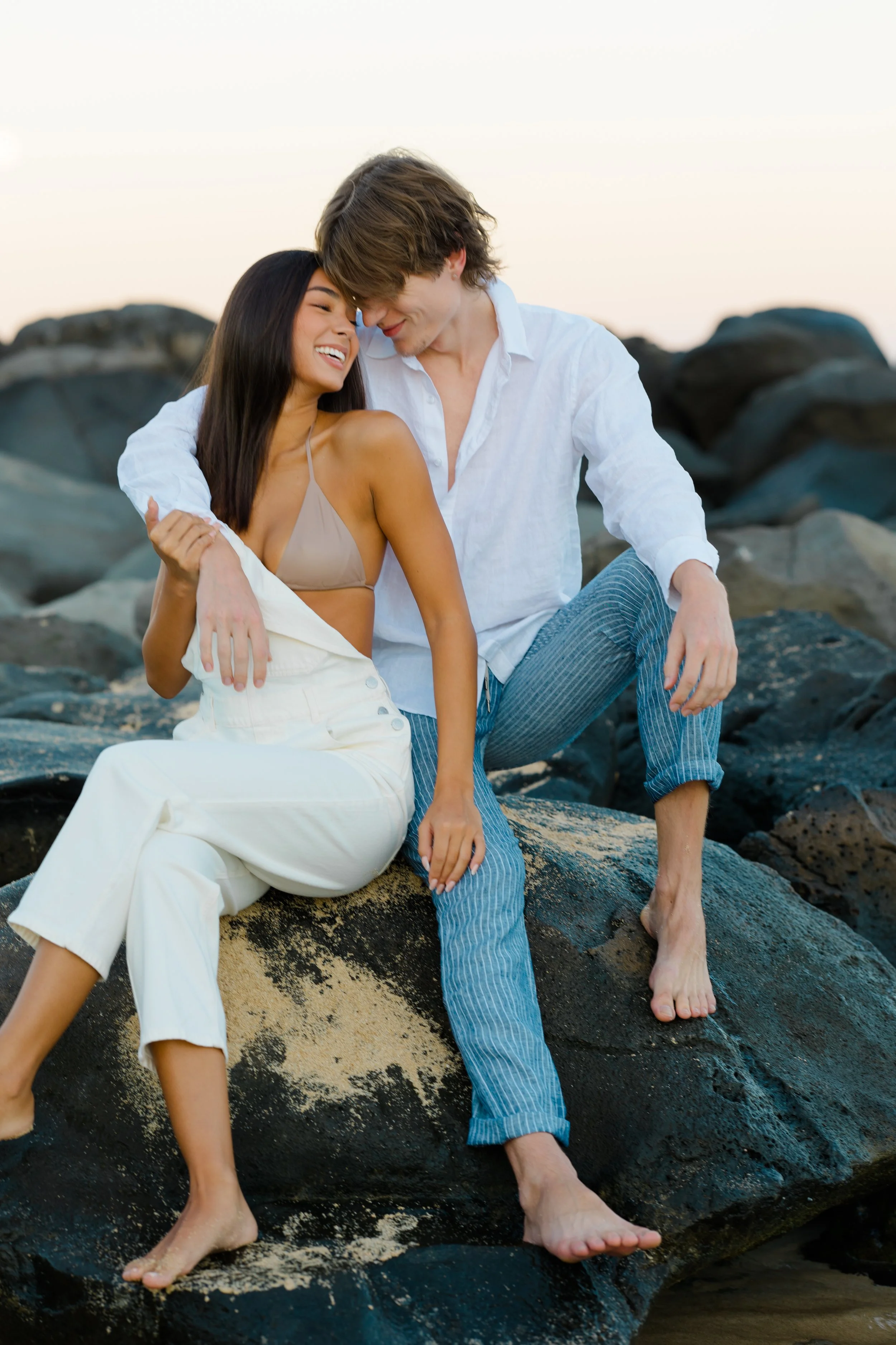 A couple sitting on rocks at the beach, smiling and leaning close to each other during sunset.