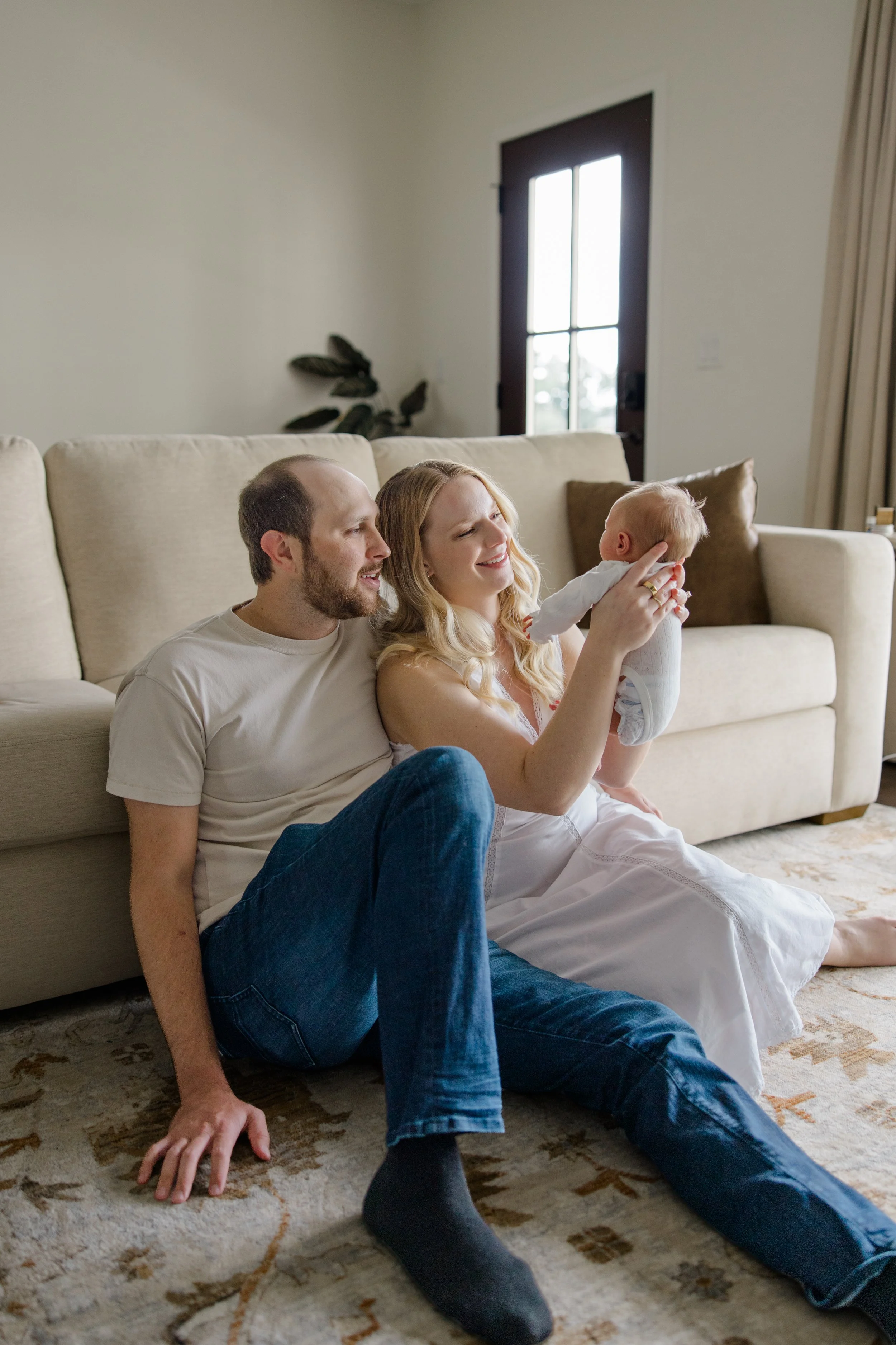 A family of three sitting on the floor in a living room, with a woman holding a baby, and a man sitting beside them, all smiling and enjoying a moment together.