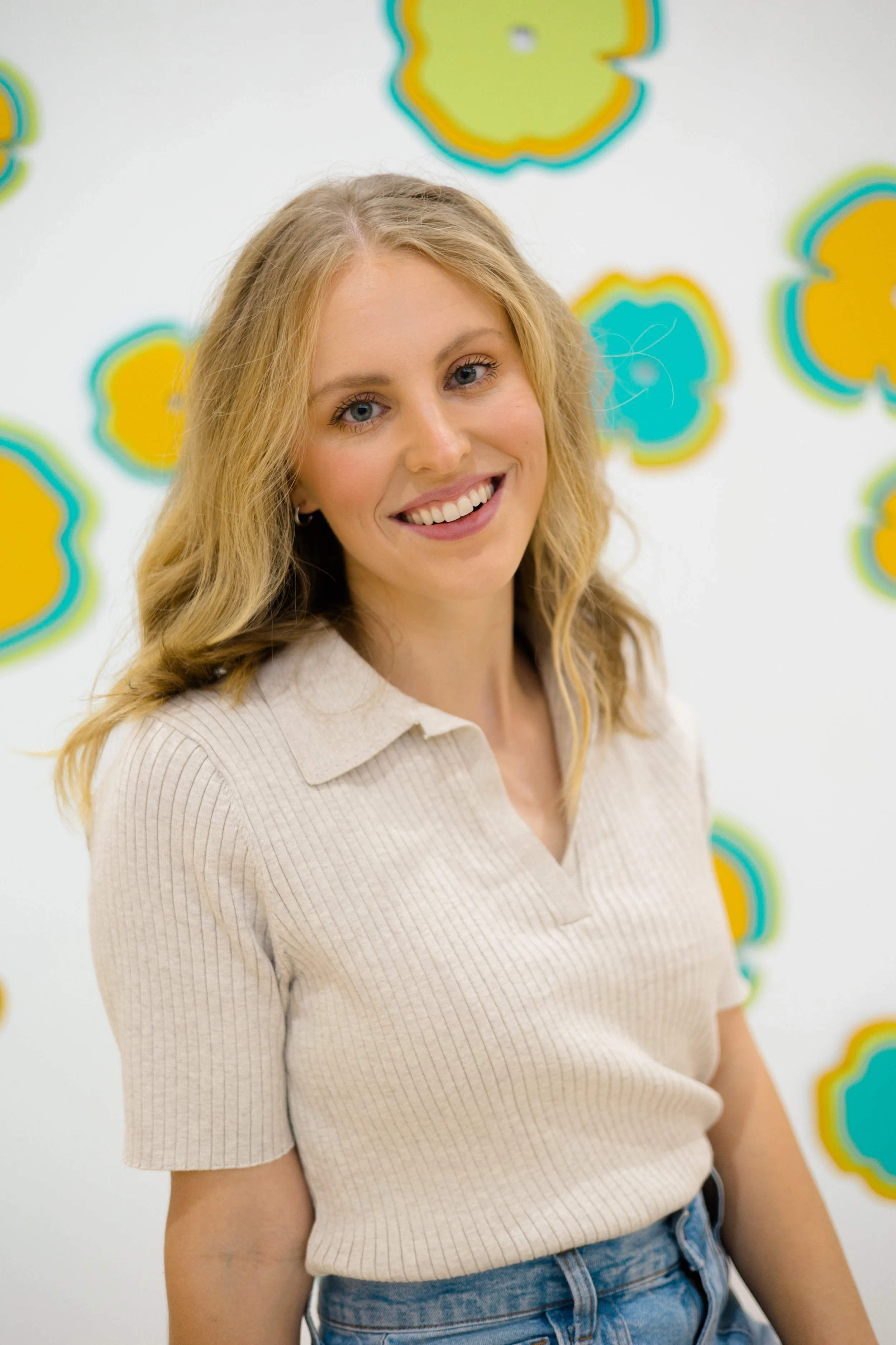 A smiling young woman with blonde hair wearing a beige ribbed short sleeve shirt and blue jeans, standing in front of a white wall decorated with colorful, outlined, irregular shapes in yellow, blue, and green.