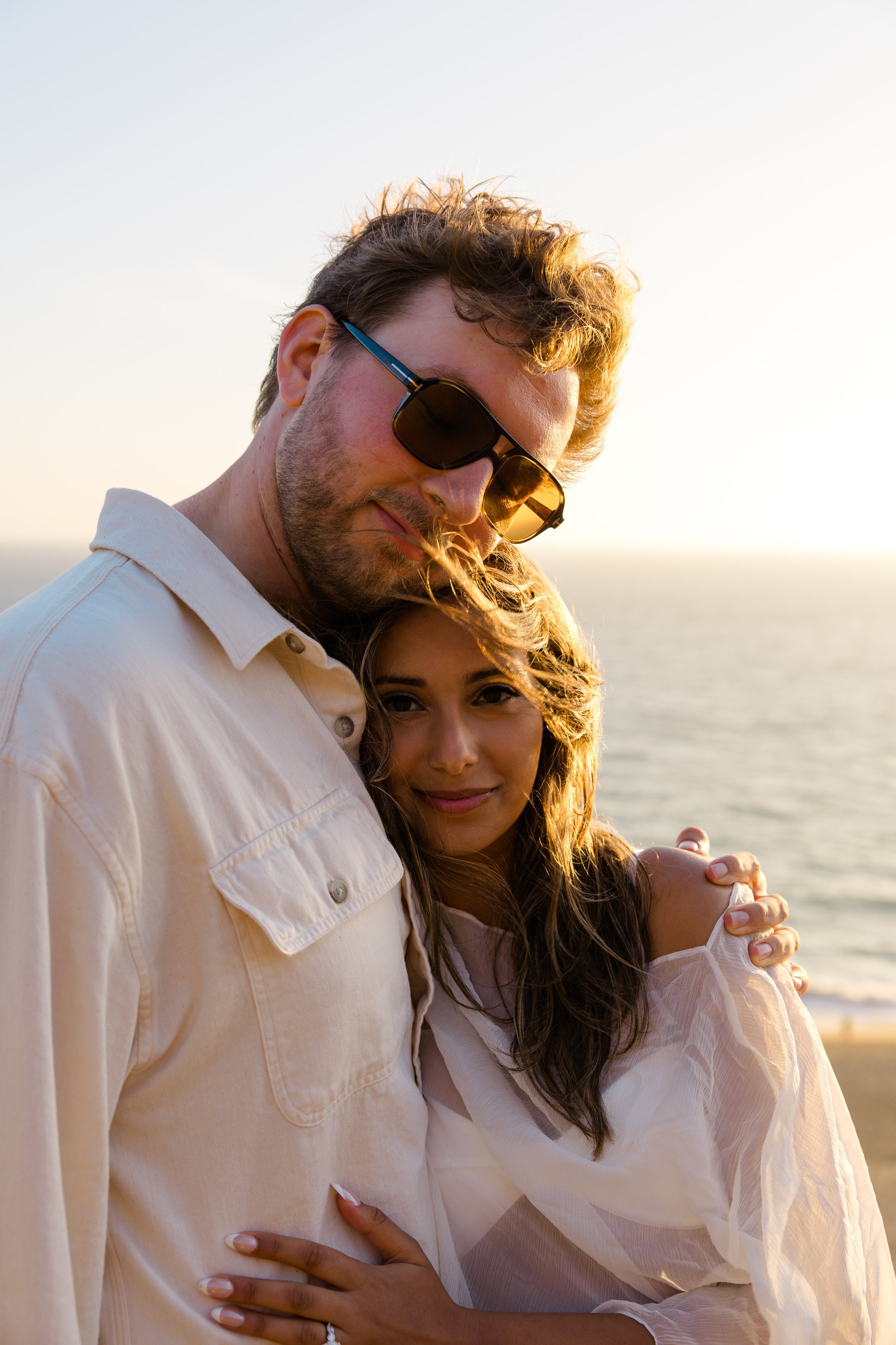 A man wearing dark sunglasses and a light-colored shirt hugging a woman, with a beach and ocean in the background during sunset.