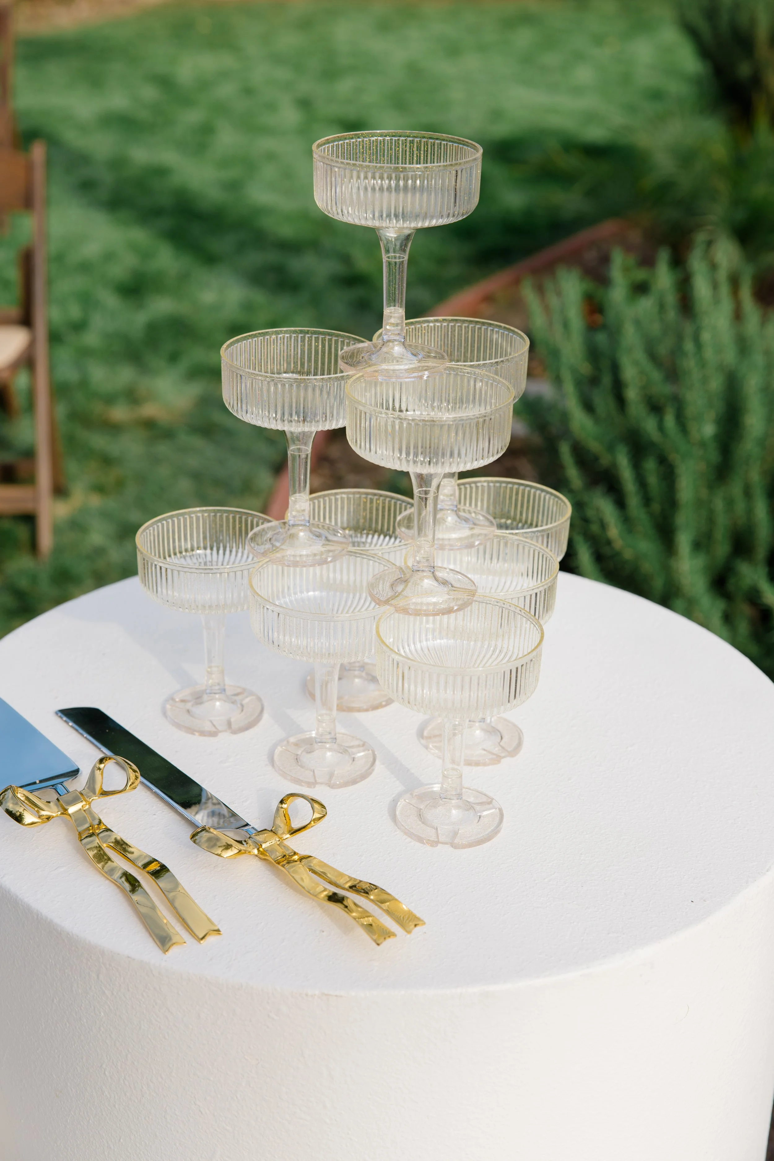 A tiered display of empty glass coupe glasses arranged on a white round table outdoors with greenery in the background, along with gold-colored scissors, a silver knife, and gold-colored tongs placed on the table.