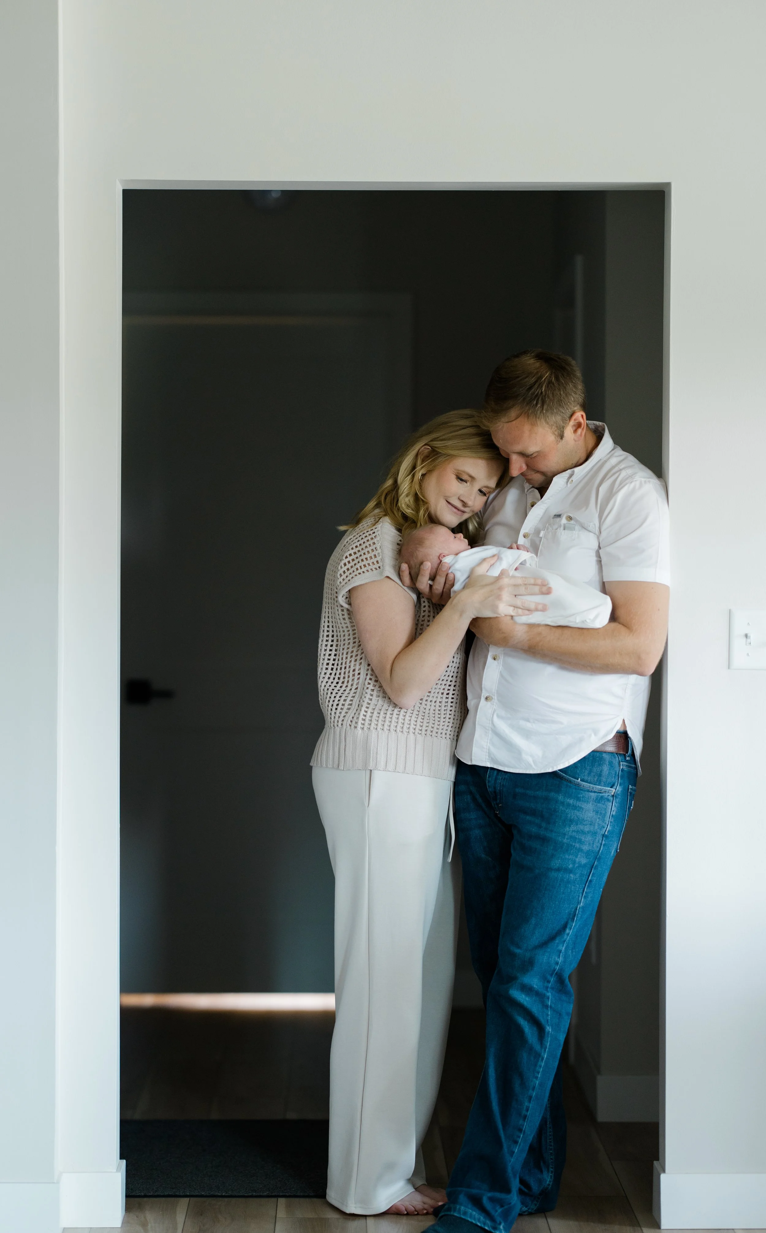 A family of three, a woman, a man, and a newborn baby, standing in a doorway in a home, smiling and holding the baby close.