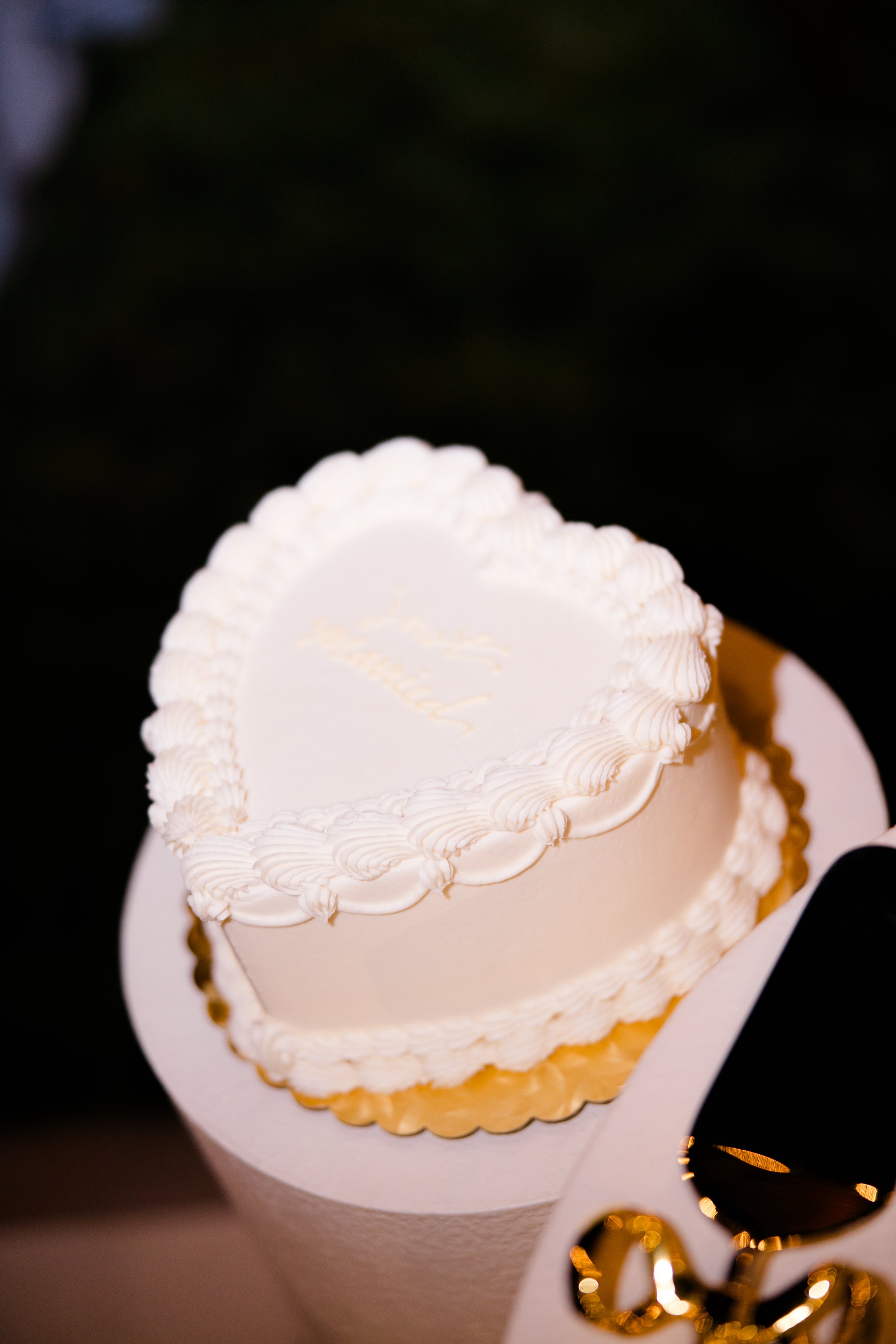 A heart-shaped cake decorated with white frosting and piped borders on top and around the sides, placed on a gold scalloped cake board.