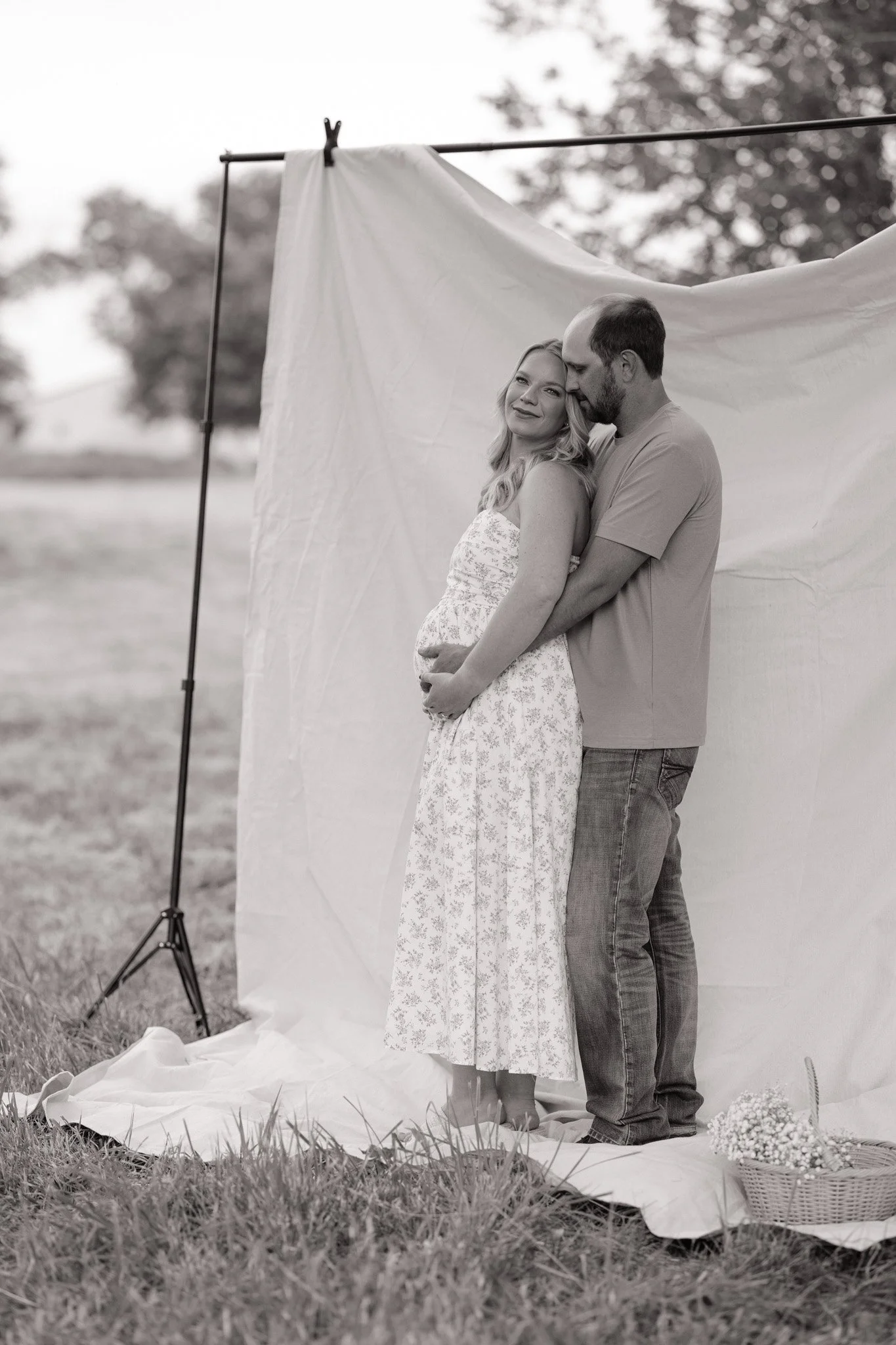 A couple standing outdoors in front of a draped white fabric backdrop, embracing and smiling. The woman is wearing a floral dress, and the man is dressed in casual clothes. There is a basket of flowers on the ground nearby. The scene appears to be a 
