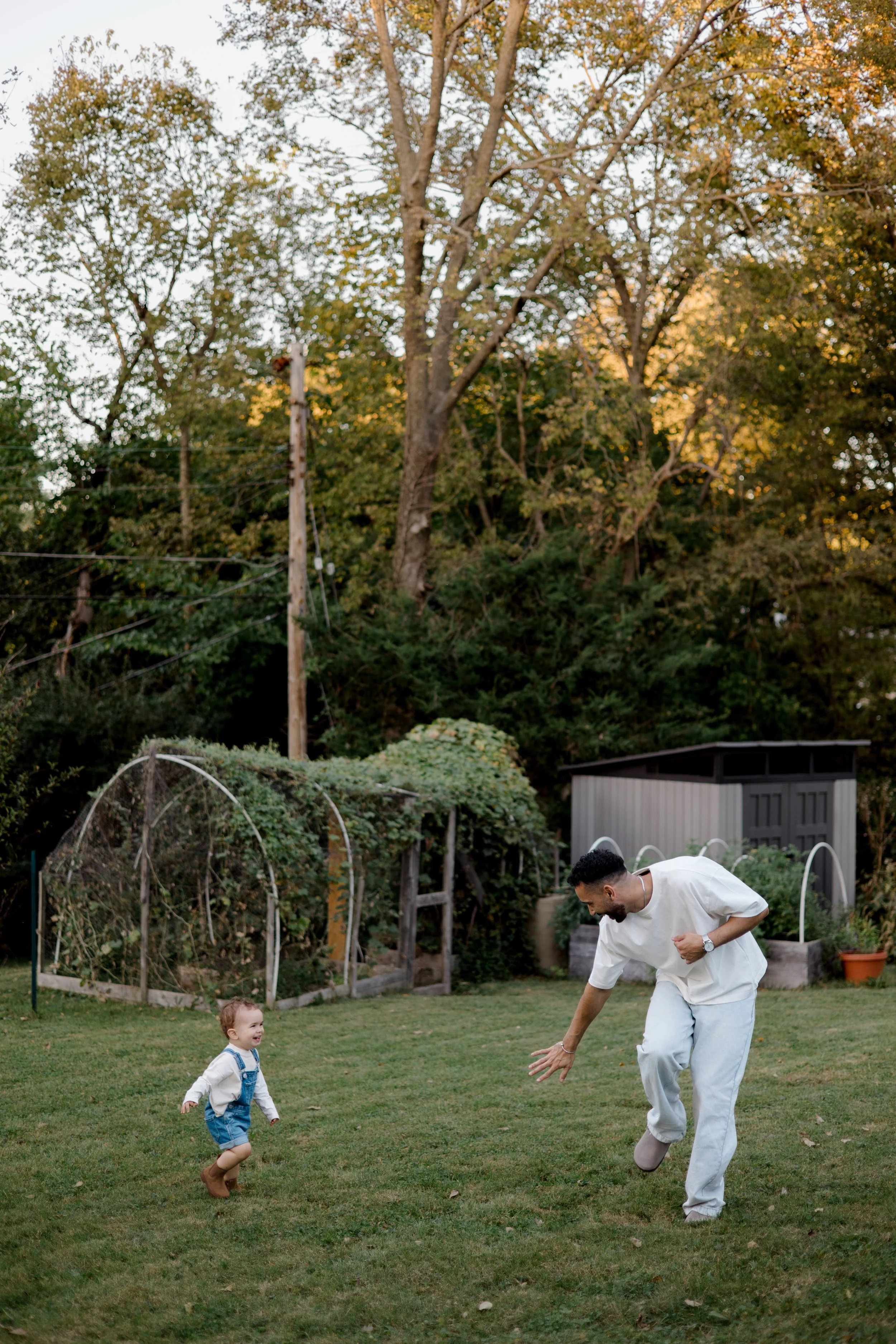 A man and a young child playing on a grassy backyard lawn during the daytime. The man is bent over, reaching towards the ground, while the child, wearing overalls and boots, is smiling and running towards him. In the background, there are trees, a ga