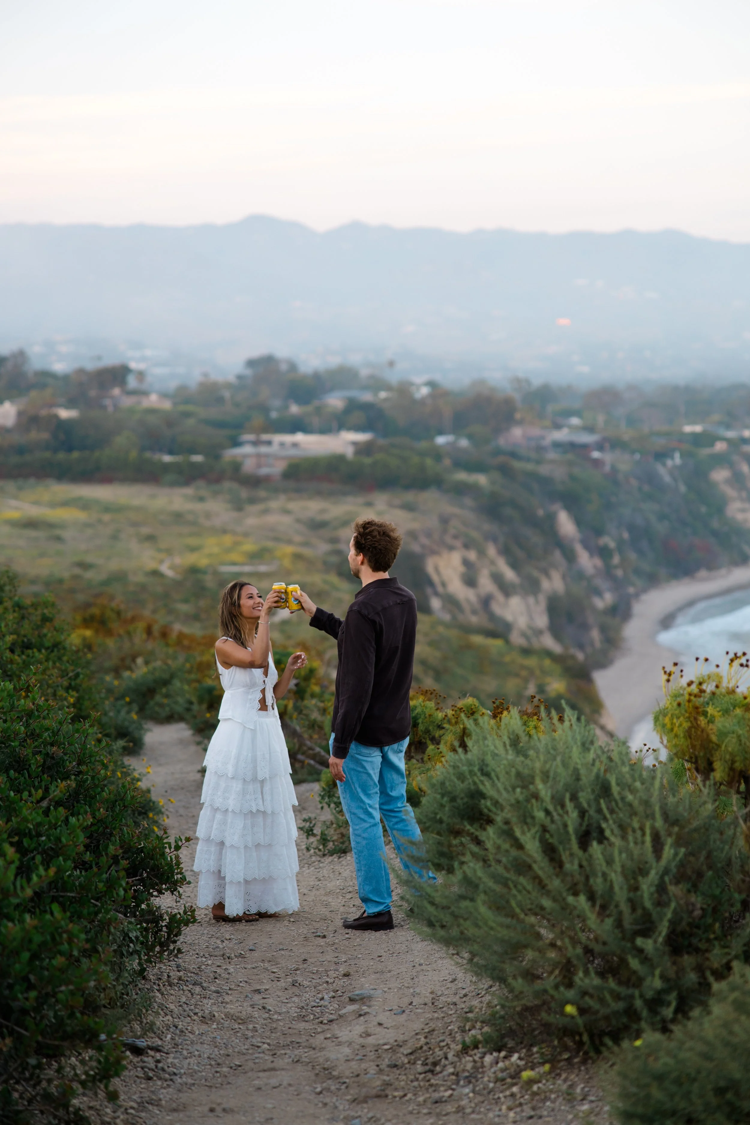 A man and woman standing on a dirt path near lush bushes, holding drinks and toasting with a scenic landscape of hills and water in the background during daytime.