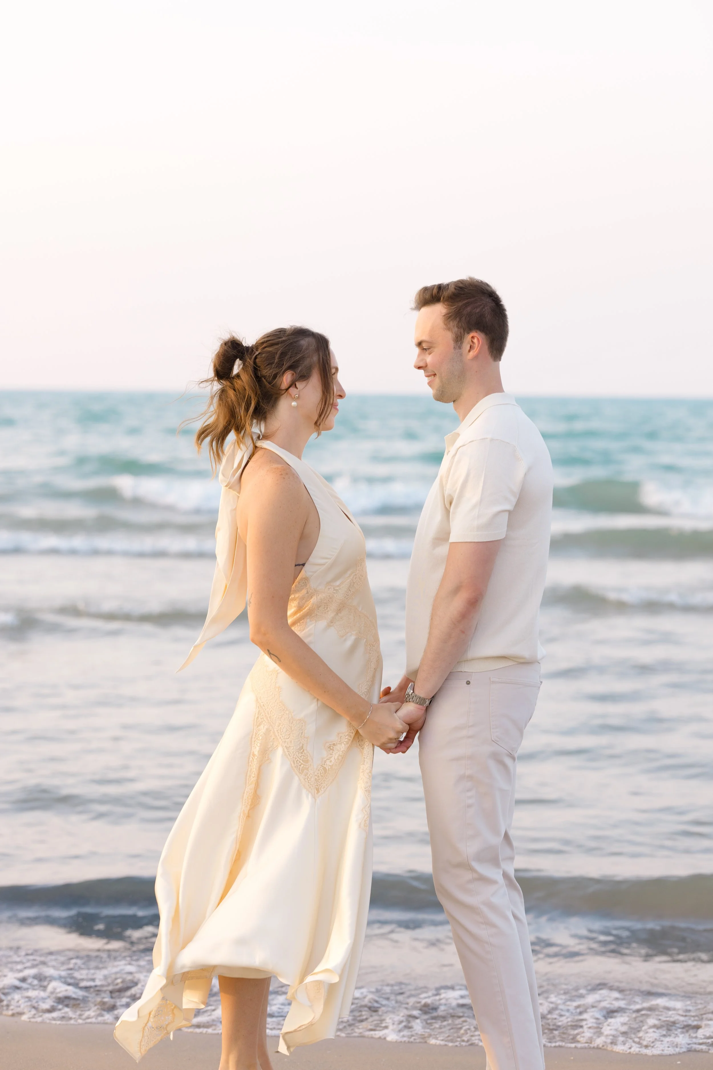 A couple holding hands on the beach, facing each other, with the ocean behind them and a light sky above.