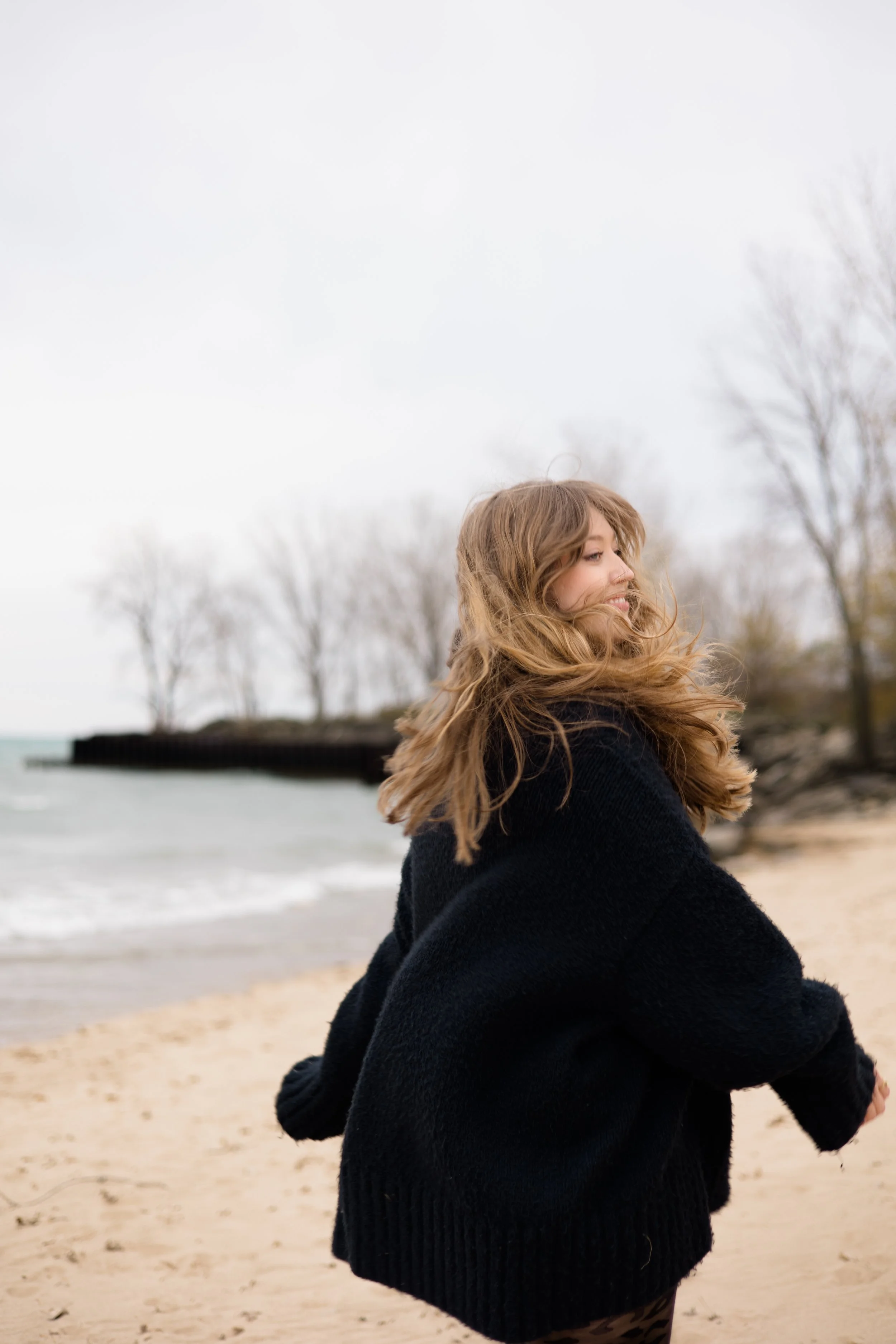 A woman with long, wavy hair is sitting on a beach with her arms crossed, wearing a black sweater, smiling, with trees and a pier in the background on an overcast day.