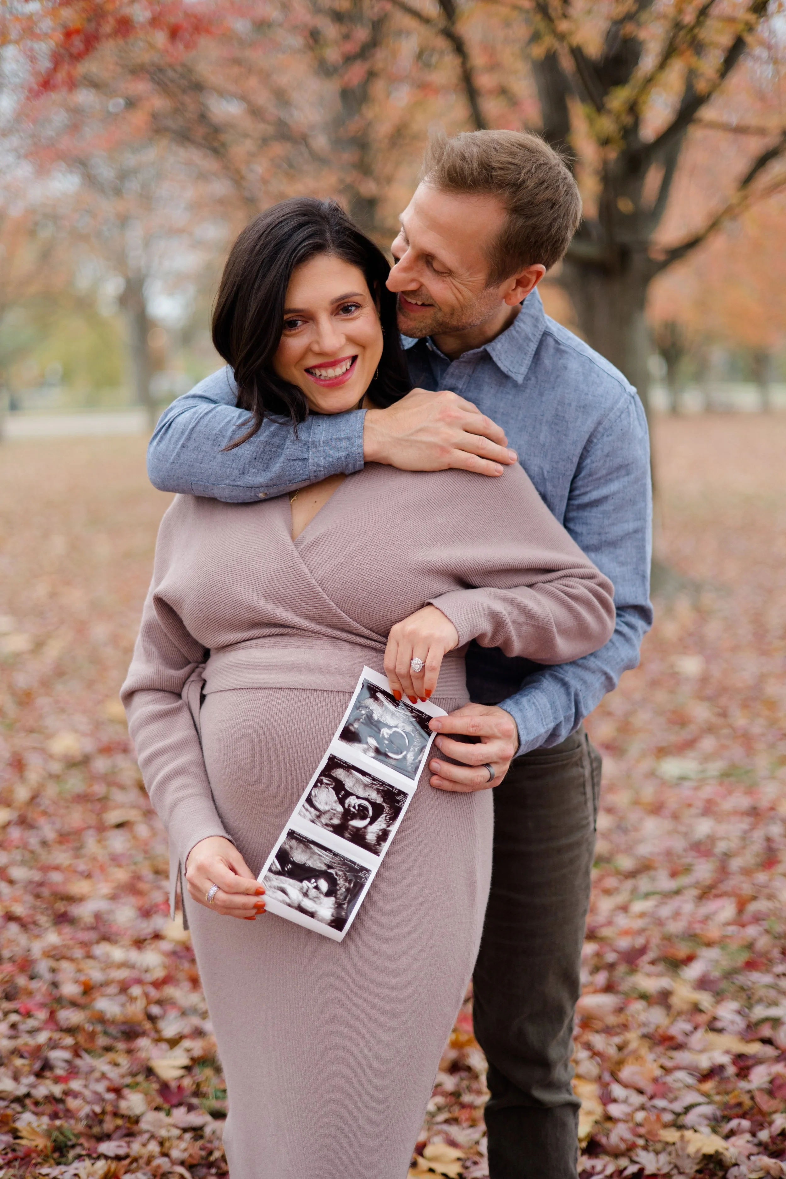 A pregnant woman holding ultrasound pictures standing in a park with autumn leaves, with a man standing beside her, smiling and looking at her affectionately.