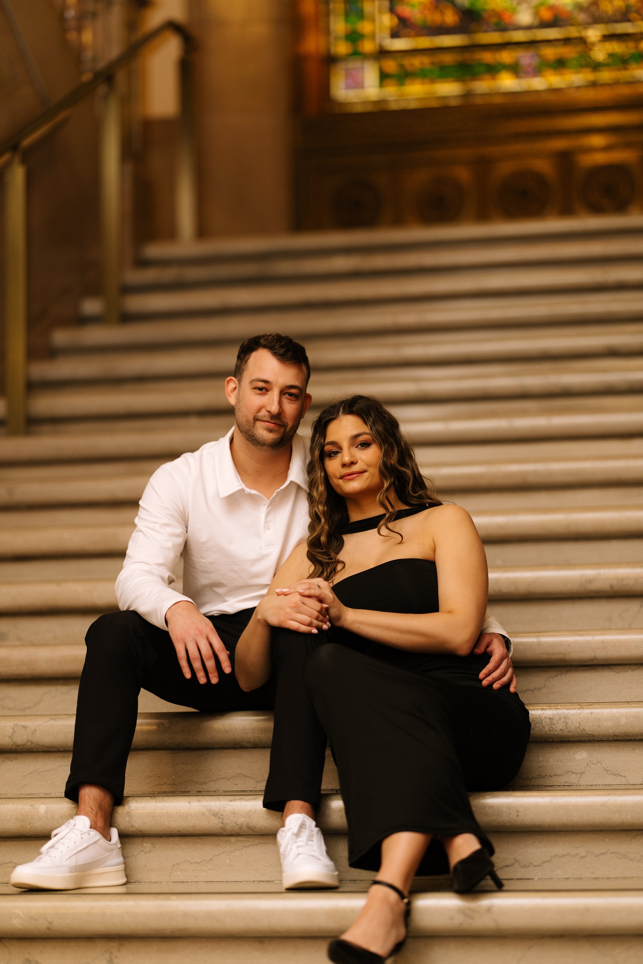 A young man and woman sitting on the stairs in an indoor setting, the man with short dark hair and a beard, wearing a white shirt and black pants, the woman with long wavy hair, wearing a black dress with an open shoulder design and black heels, posi