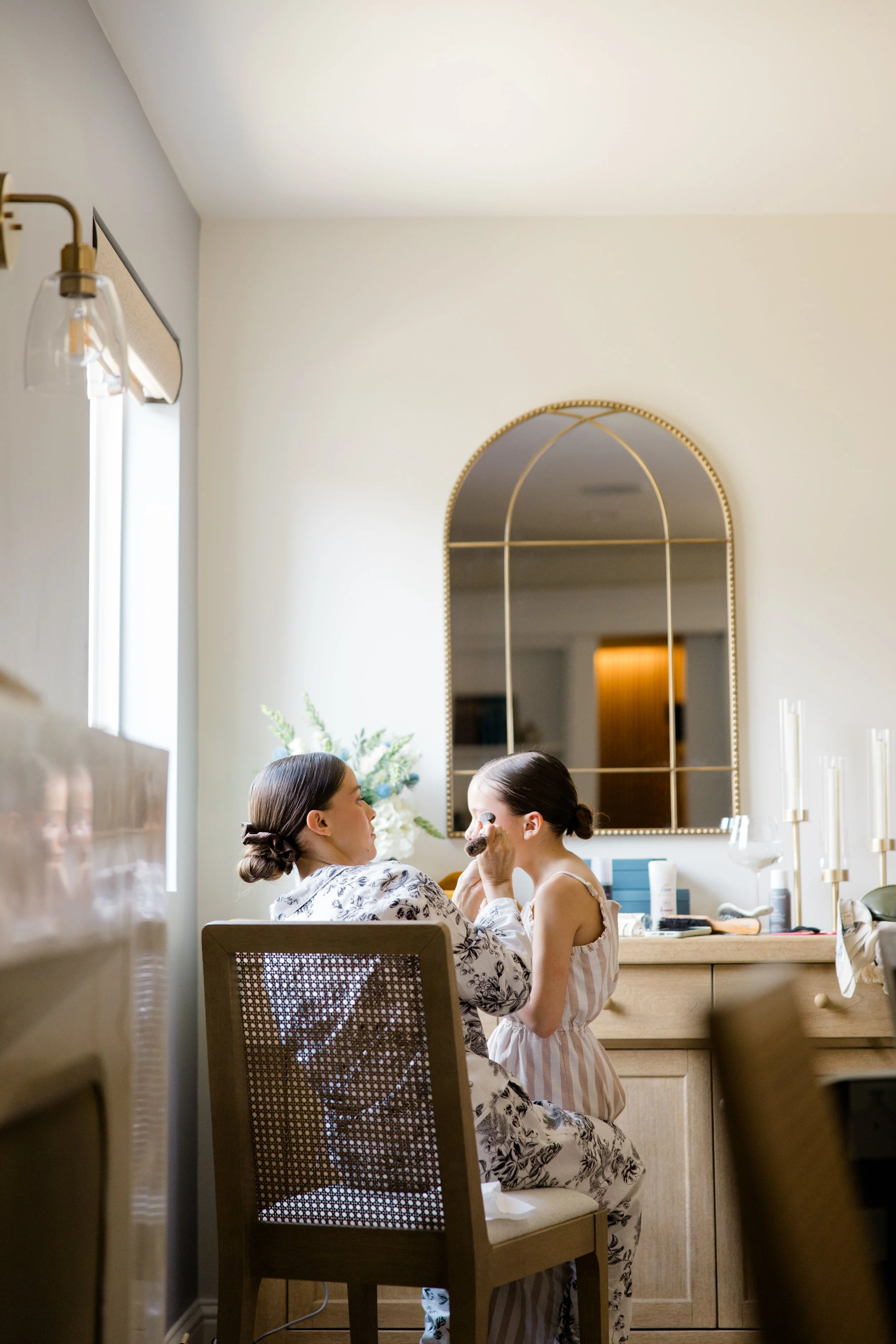 A woman is applying makeup to a young girl in a cozy, well-lit room with a large mirror, wall-mounted light fixtures, and decorative elements.
